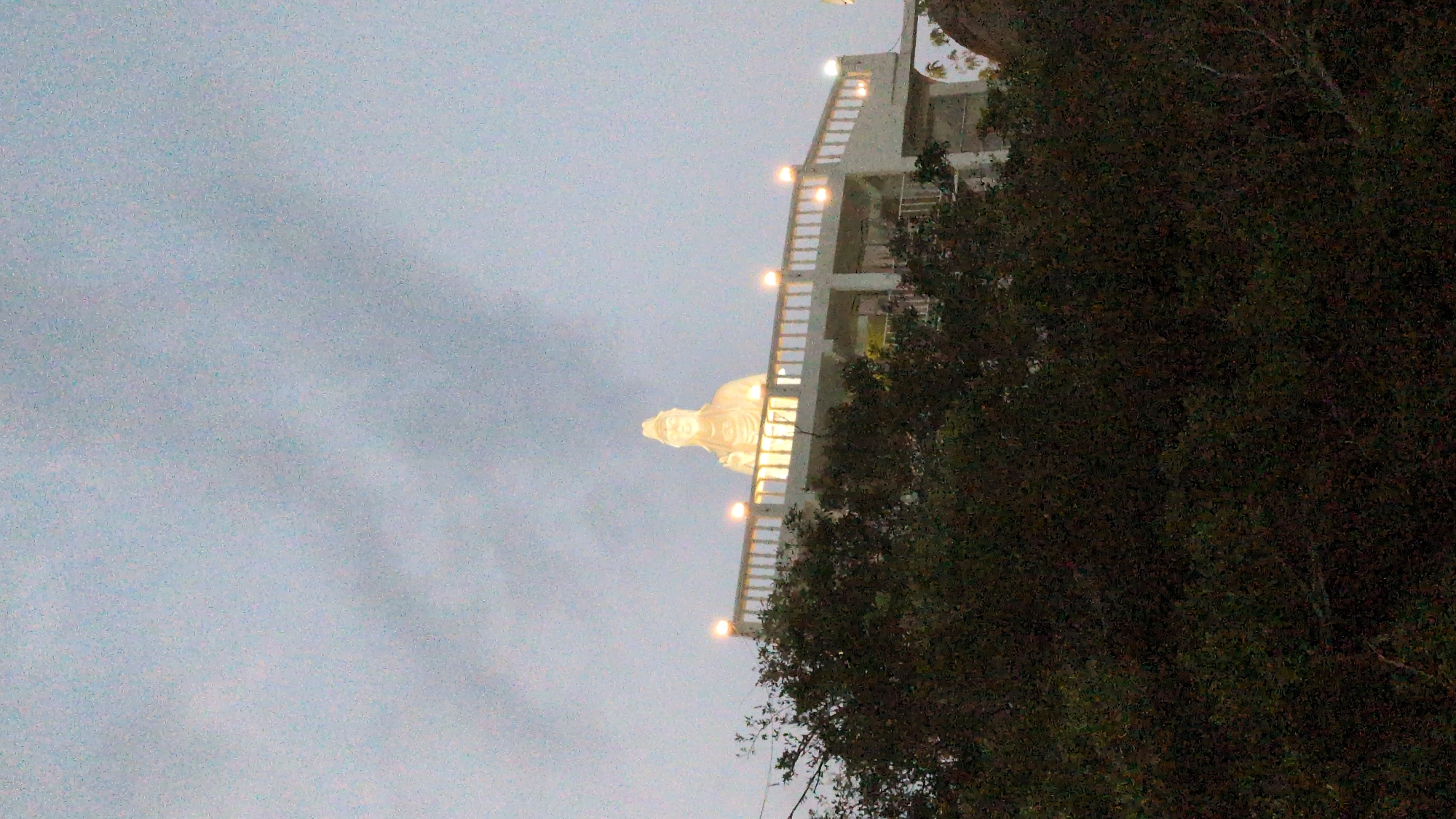 A medium-distance shot shows the Guan Yin statue atop a staircase-like structure, illuminated with lights in Khánh Hải, Vietnam. The statue is partially obscured by trees on the right side. The left side of the frame shows a cloudy, dusky sky.