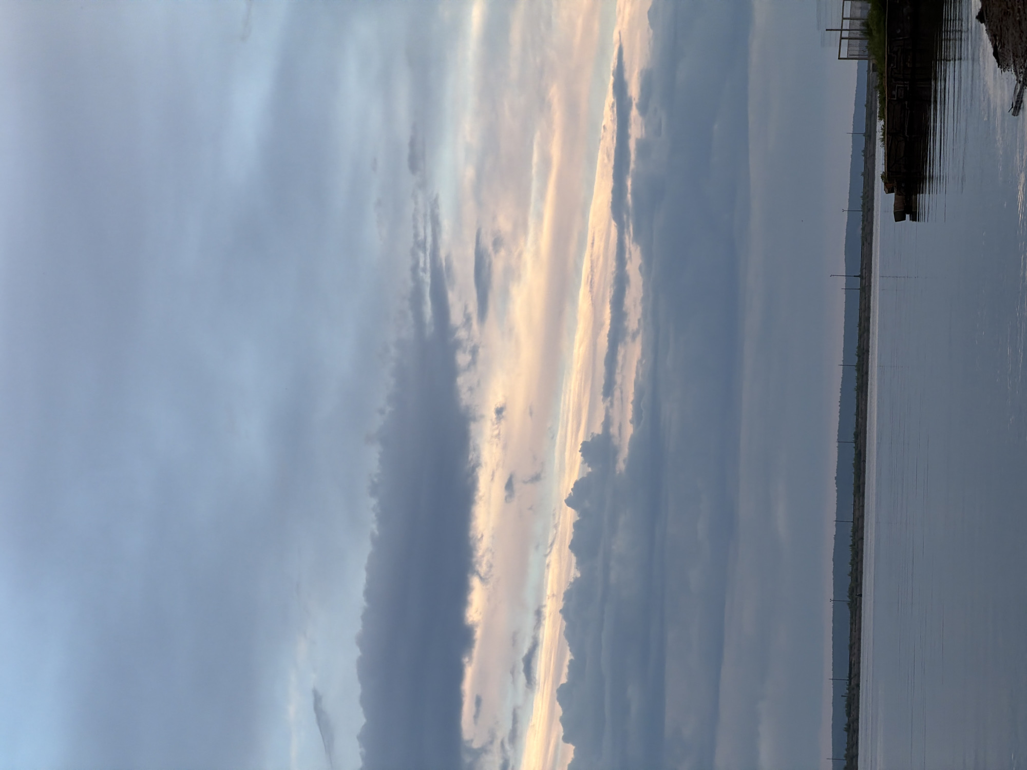 The image depicts a twilight scene over the Kama River in Chaikovsky, Russia. The sky is filled with clouds, ranging from dark gray to light pink and white hues. The river is calm, reflecting the colors of the sky. Along the riverbank, there is a pier or dock structure. Utility poles can be seen in the background along the shoreline.