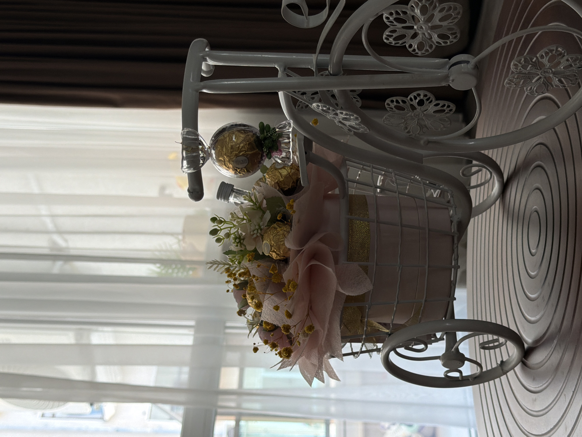 A decorative white metal bicycle holds a floral arrangement and chocolates. The bicycle is positioned on a circular wooden surface. In the background, sheer white curtains are visible, with a darker brown curtain to the right. The scene appears to be indoors, near a window, and is not located in Veliko Tarnovo, Bulgaria. There are no people or actions depicted in the image.