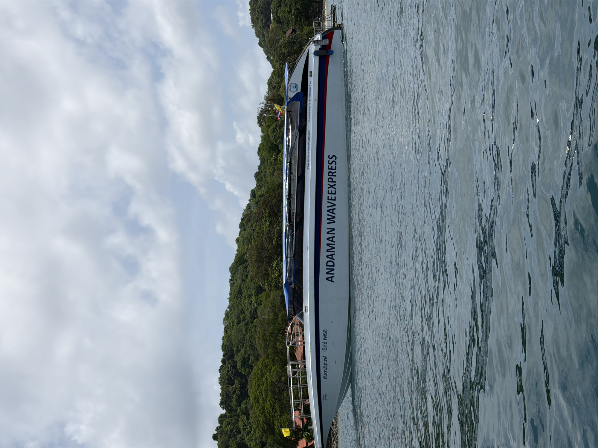 A speed boat, named "ANDAMAN WAVEEXPRESS," is docked in the water near a lush, green, forested island, under a cloudy sky. The boat is white with blue and red stripes. The island features trees and some buildings, including what appears to be a resort with red-tiled roofs. The water is calm with ripples, reflecting the sky and surrounding environment. The scene is tranquil and depicts a typical view of the Andaman coast near Ao Nang, Thailand, a popular tourist destination known for its stunning natural beauty and island hopping activities.