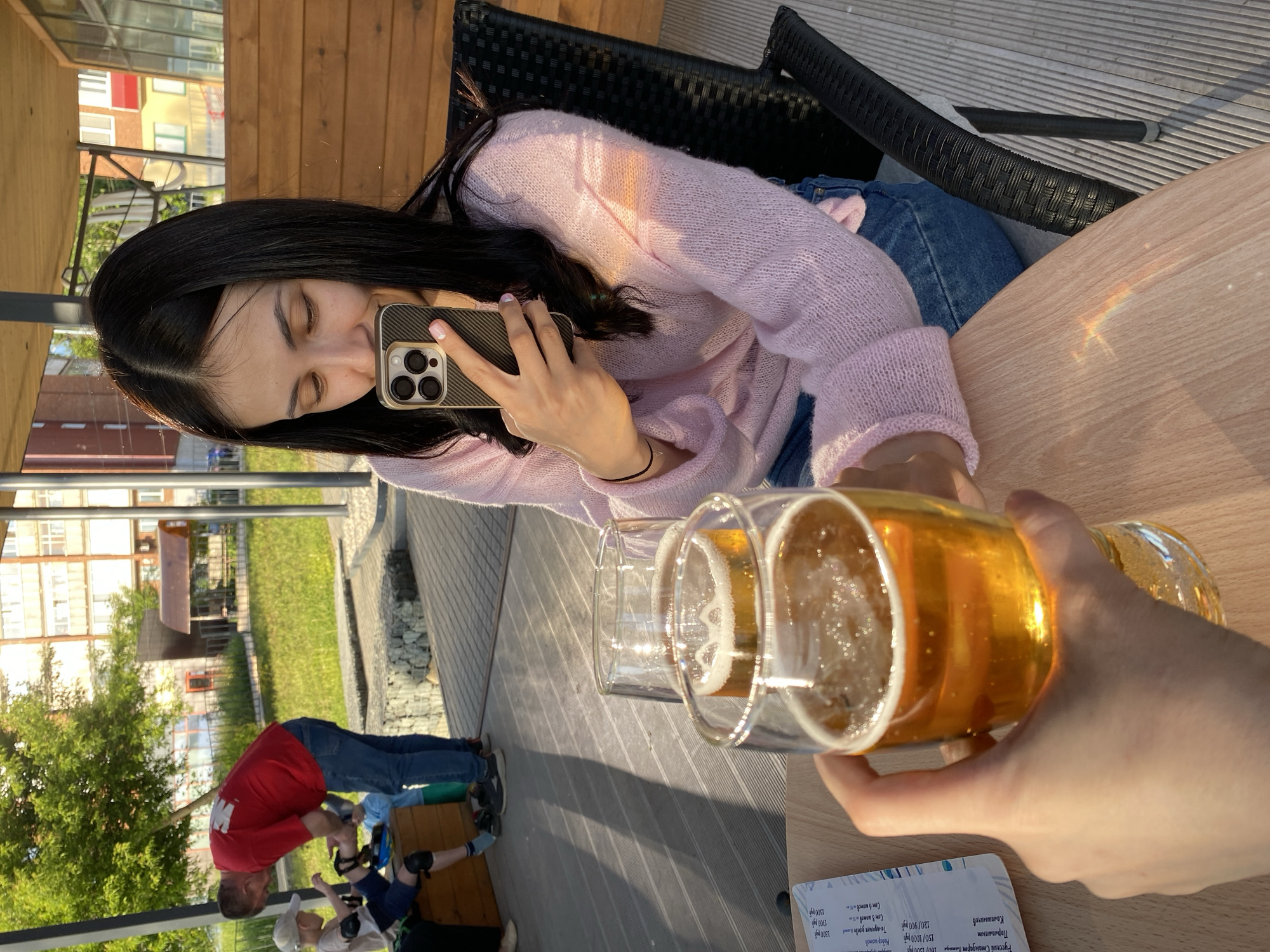 The image captures a casual outdoor scene, likely at a cafe or patio in Votkinsk, Russia, during the late afternoon or early evening, indicated by the warm, directional sunlight and long shadows. A young woman with long, dark hair is the central focus, seated at a light wooden table. She is wearing a light pink, possibly knitted, sweater and blue jeans. Her eyes are downcast or closed as she intently looks at her smartphone, which has a textured dark case, held in her left hand. Her nails are painted light pink.
In the immediate foreground, a hand, likely the photographer's, holds a glass of golden beer with ice and foam. Another similar glass is visible on the table. A small, faint rainbow-colored light refraction is visible on the table surface near the woman's leg. The seating area features dark woven chairs and a wooden deck.
In the background, beyond the patio, there's a grassy area with green trees and distant multi-story buildings, some with yellow and red facades. A man in a red t-shirt and blue jeans is seen bending over, interacting with a small child who wears a light-colored hat or helmet and knee/elbow pads, suggesting outdoor play.
A menu or price list is visible on the table, featuring text such as "Русский Стандарт Платинум 100/1500/90", "Парламент", "Калашников", and various prices like "150/1000 руб Набор шотов", "120/900 руб Танауиющие дерева 3500 قوم", "1700 руб", and "1300 руб", indicating a selection of alcoholic beverages. The overall mood is relaxed and leisurely.