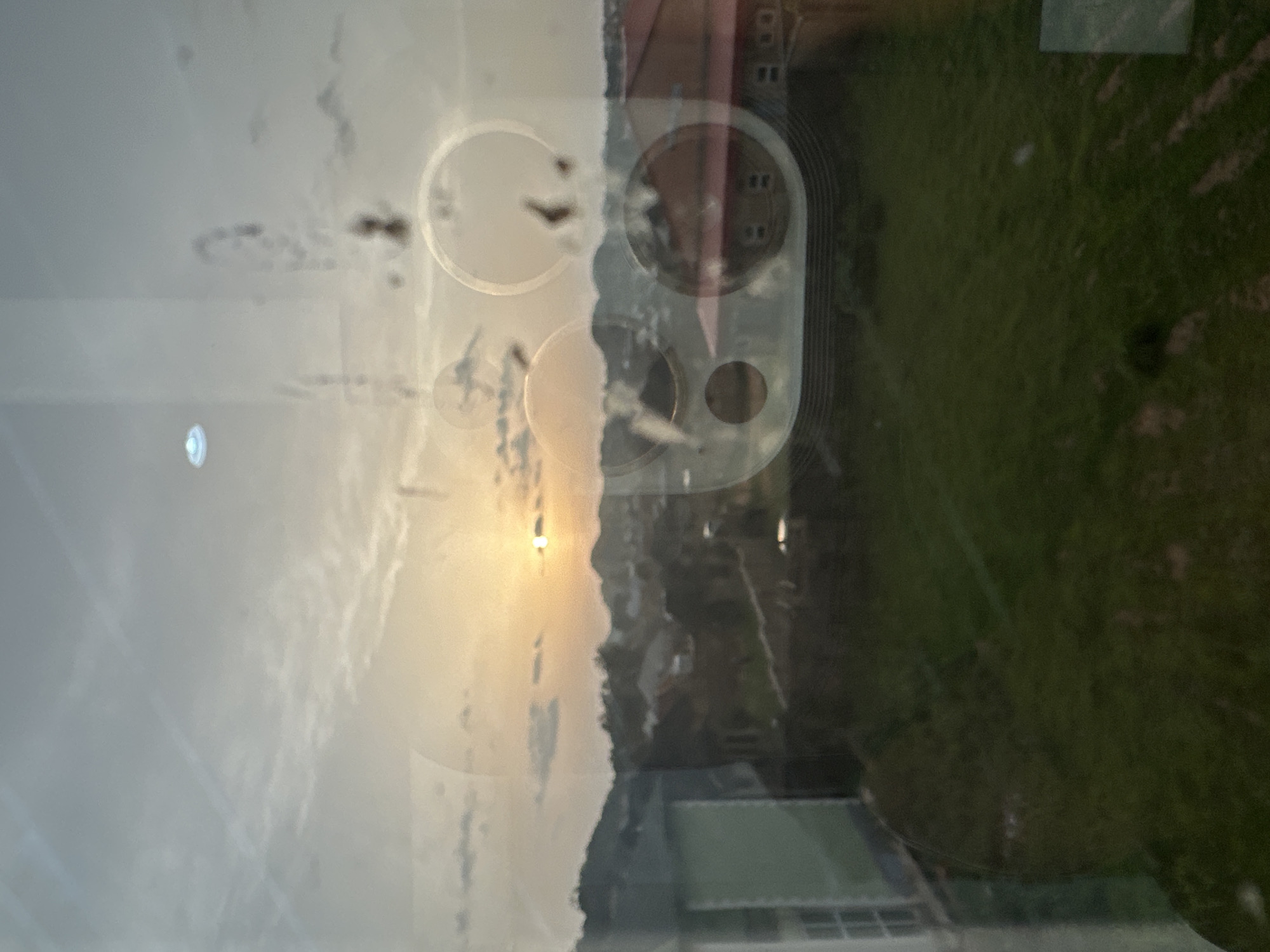 The image shows a hazy view through a window, partially obscured by circular smudges and a phone's camera module reflection. Outside the window is a landscape with a pale sky, possibly at sunset, and a view of a building and green vegetation. There's no evidence to support this is in Bukuru, Nigeria. There are no people visible.
