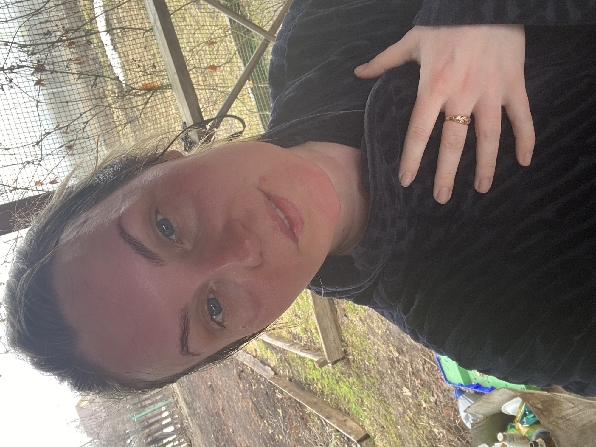 A woman with fair skin and blue eyes poses for a selfie. She is wearing a dark blue, textured top and a gold ring on her right ring finger. Her face is slightly flushed, and she has minimal makeup. The background suggests an outdoor setting, possibly a garden or a yard, with a fence-like structure and bare branches visible. The ground has fallen leaves and some green vegetation. There are also some containers and other objects scattered around in the background. Based on information from online sources, it is possible that this location is near Chaikovsky, Russia, although specific landmarks are not visible.