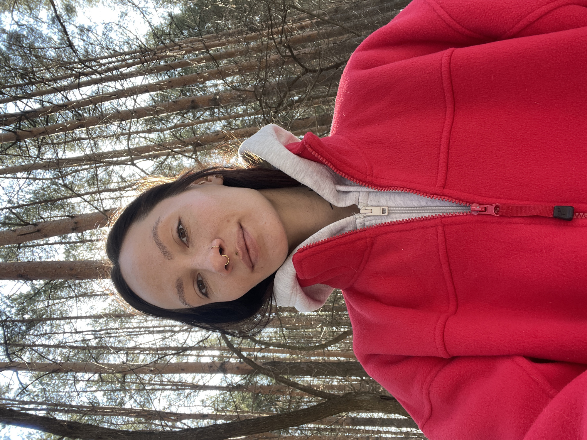 A woman is in a forest, looking at the camera. She wears a red fleece jacket, with a gray hoodie underneath. Bare trees rise up behind her. There is no verifiable information to suggest this photo was taken in Izhevsk, Russia.