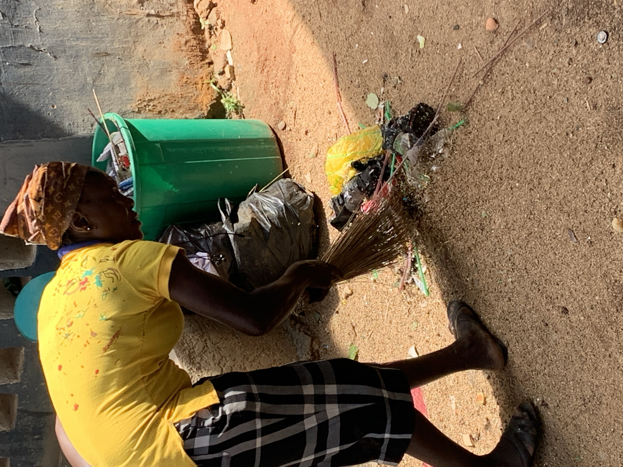 A person wearing a yellow shirt, black and white checkered skirt, and a brown headscarf is sweeping up trash with a straw broom. The person is barefoot. The trash includes plastic bags (yellow and black), straws, and general debris. A green trash can is positioned nearby, partially filled with trash. The location appears to be an outdoor area with a sandy or dirt surface. The location is New Stadium, Nigeria.