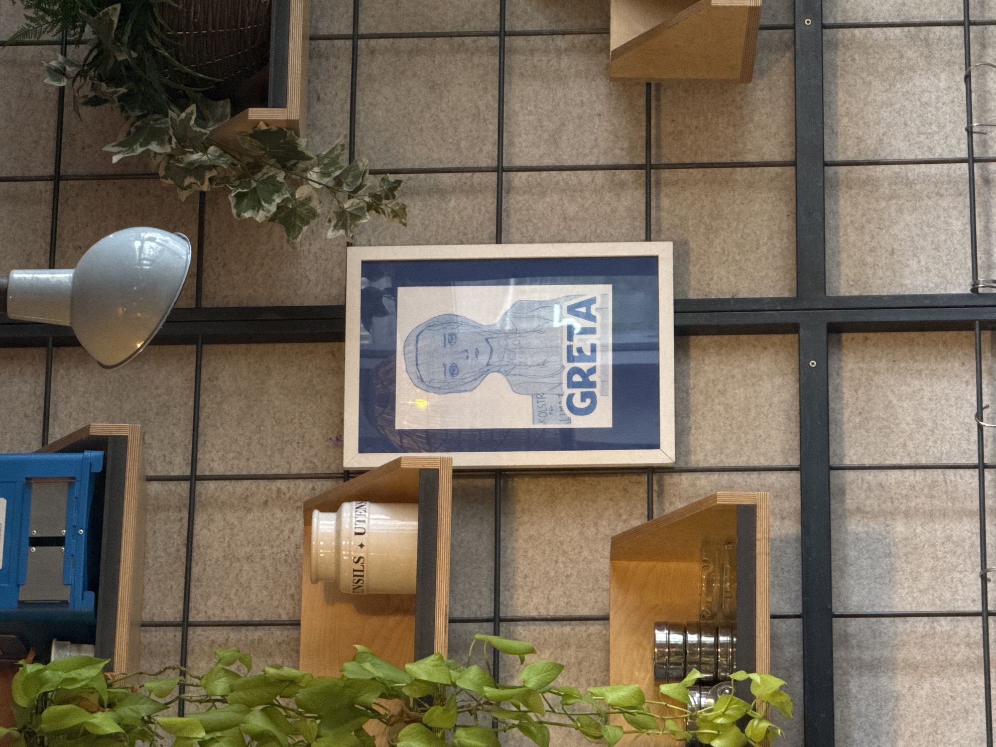 The image shows a section of a wall decor in what appears to be a commercial or public space, possibly a café or shop, within the city of London, United Kingdom. The wall is covered with a grid of dark metal bars. On the wall are displayed various decorative elements and storage units made of wood.
Among the decorative elements is a framed print of Greta Thunberg, with her name prominently displayed, adding a political or social commentary element to the décor. Additionally, there is a white ceramic jar with “ENSILS + UTEN” printed on it, sitting on a small wooden shelf. A blue metal box is situated on another shelf on the left side of the picture.
Green plants are strategically placed to add a natural touch to the industrial grid design of the wall. A silver lamp is fixed to the wall, reflecting light. The image suggests a contemporary and slightly eclectic interior design.