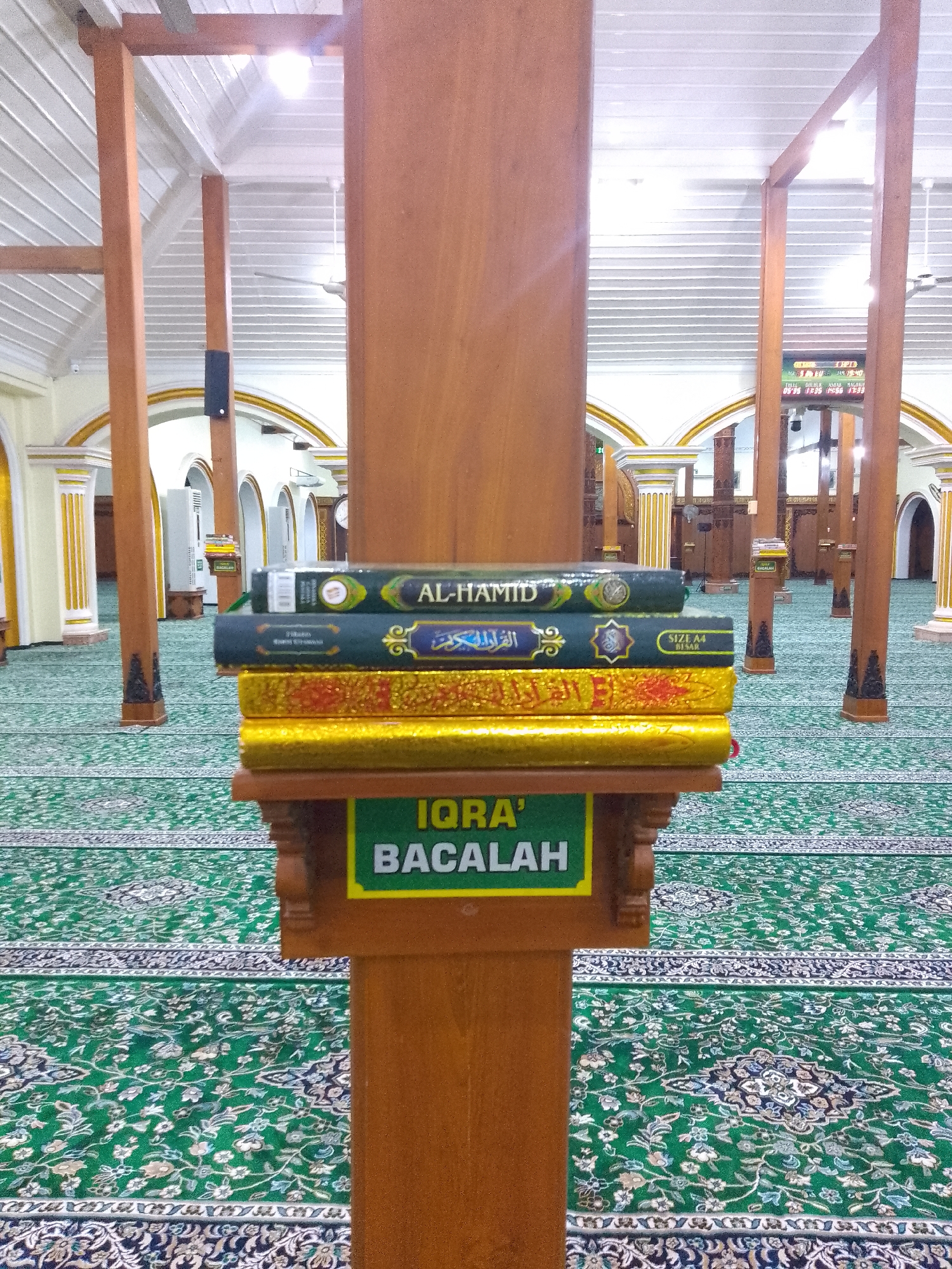 A stack of four books rests on a wooden display stand in the center of a carpeted hall. The books, bound in green and gold, are arranged horizontally. The top book has "AL-HAMID" written on its spine in white lettering. Below it, another green book has Arabic script. The bottom two books are gold. The display stand has a green sign that reads "IQRA' BACALAH" in white letters. The hall, which appears to be a mosque, has a green carpet with intricate patterns and tall wooden pillars supporting a white, slatted ceiling. Natural light and ceiling lights illuminate the space. In the background, rows of similar wooden pillars and arched doorways can be seen. This image was taken at Masjid Agung Lamongan in Lamongan, Indonesia.