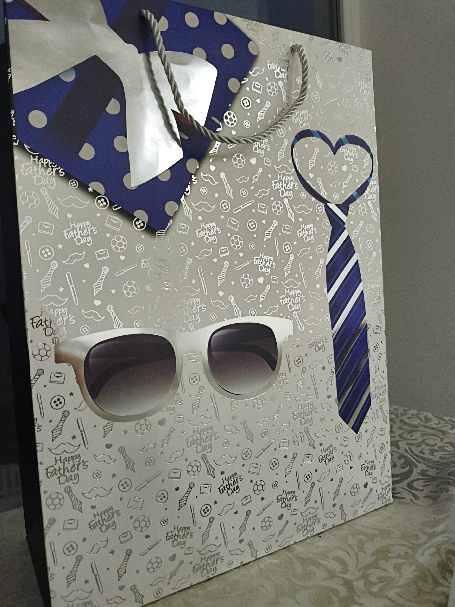 A Father's Day gift bag features a pattern of small icons, including mustaches, ties, soccer balls, briefcases, pens, and buttons, interspersed with the phrase "Happy Father's Day". The gift bag is designed to resemble a face, with a blue and white striped tie as a mouth, and white-rimmed sunglasses serving as eyes. A blue bow tie with white polka dots is positioned at the top, resembling a head. The gift bag rests on a textured surface, possibly a table, with a blurred background. The scene is captured in an indoor setting.