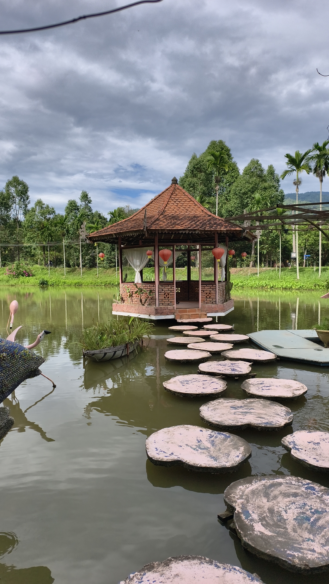 A serene lake with a central gazebo and a path of stepping stones occupies the foreground of this outdoor scene. The gazebo, constructed of red brick and featuring a multi-sided, reddish-brown tiled roof, is positioned on a raised platform at the lake's edge. White curtains hang from the gazebo's openings, and several red lanterns are suspended from the eaves. The stepping stones, irregularly shaped and varying in color from light pink to grey, are arranged in a winding path across the water, leading from the shore to the gazebo. In the left foreground, a decorative flamingo sculpture is partially visible. The lake's surface is calm and reflects the overcast sky and surrounding greenery. The background consists of lush green trees, including palm trees, and a distant mountainous landscape under a cloudy sky. The environment appears to be a park or garden setting. There are no people or animals other than the decorative flamingo.