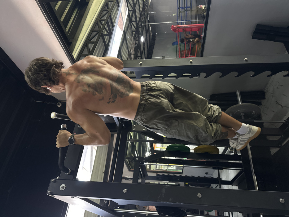 The image depicts the interior of a gym in Tibubeneng, Indonesia. A shirtless male, with brown wavy hair and a large dark tattoo covering his upper back, is performing a pull-up on a black metal power rack's pull-up bar. He wears green/khaki elastic-waist cargo capri pants, white socks, and white Adidas sneakers with beige soles. A black smartwatch is visible on his left wrist, and white wireless earbuds are in both ears. His back is to the camera, and his body is elevated from the floor, hands gripping the bar.
In the background, behind the primary subject and the power rack, a reflective glass partition reveals an adjacent area of the gym. This reflection shows a boxing ring with red and blue ropes. Inside the boxing ring, a person is seated on the floor, wearing dark attire. Other gym equipment, including weight plates (green, yellow) on a rack and a barbell, are partially visible in the mid-ground and reflected areas. The gym features a black ceiling with exposed metal structural elements and a light-colored floor.
