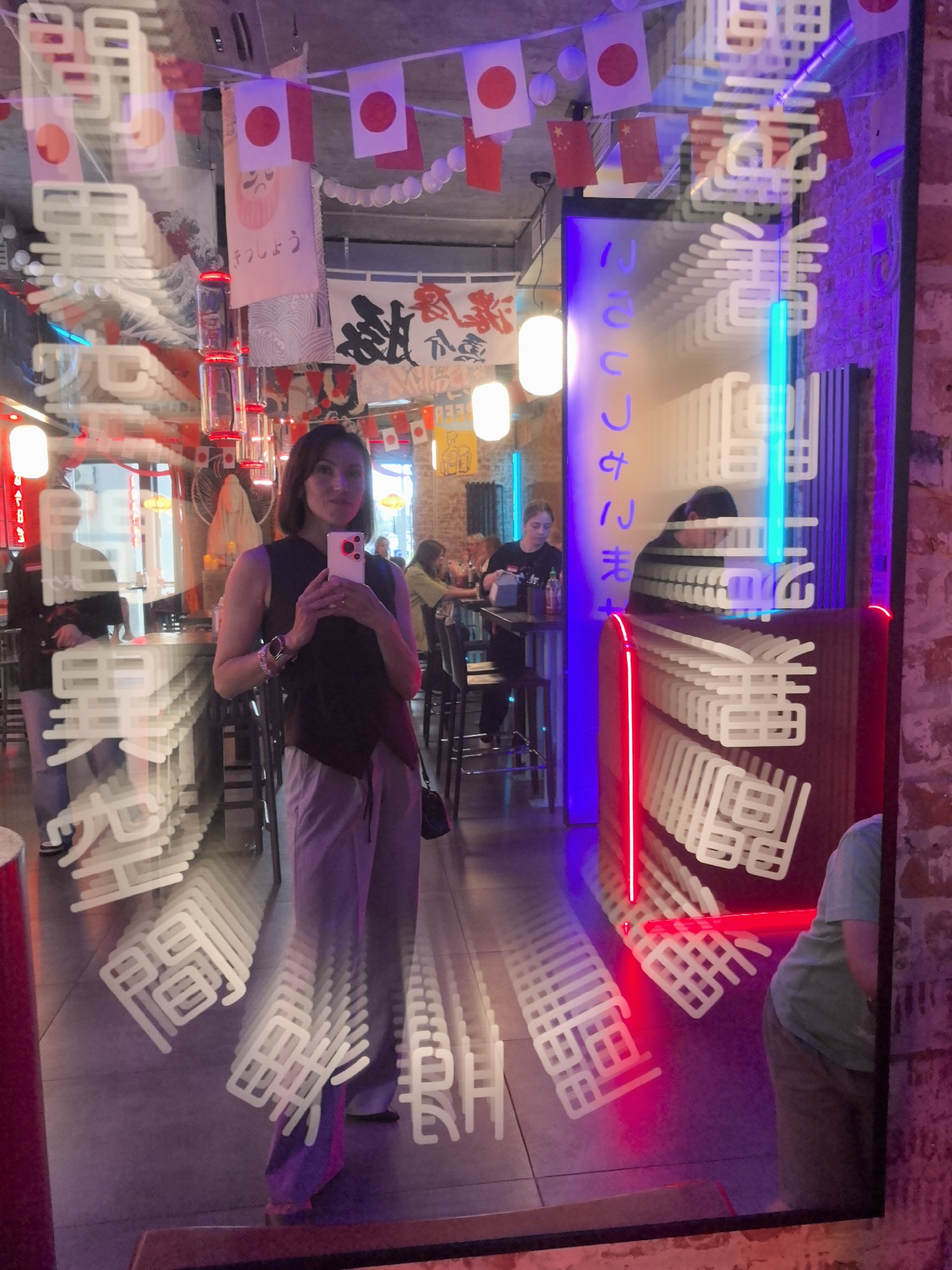 The image captures a reflection in a large mirror within a Japanese-themed restaurant. The restaurant appears to be located in the city of Perm, Russia, suggested by the presence of Japanese flags and signage, common decor in such establishments. In the foreground of the reflection, a woman stands, holding a smartphone and taking a selfie. She is wearing a dark, sleeveless top and light-colored pants. The mirror reflects the interior of the restaurant, which features a bar area, tables, and patrons. The decor includes Japanese banners, red and white string lights, and neon accents, creating a vibrant atmosphere. The woman is positioned centrally in the reflection, and her image is distorted by the reflective surface, creating multiple overlapping reflections. The background shows other patrons and the general ambiance of the restaurant.