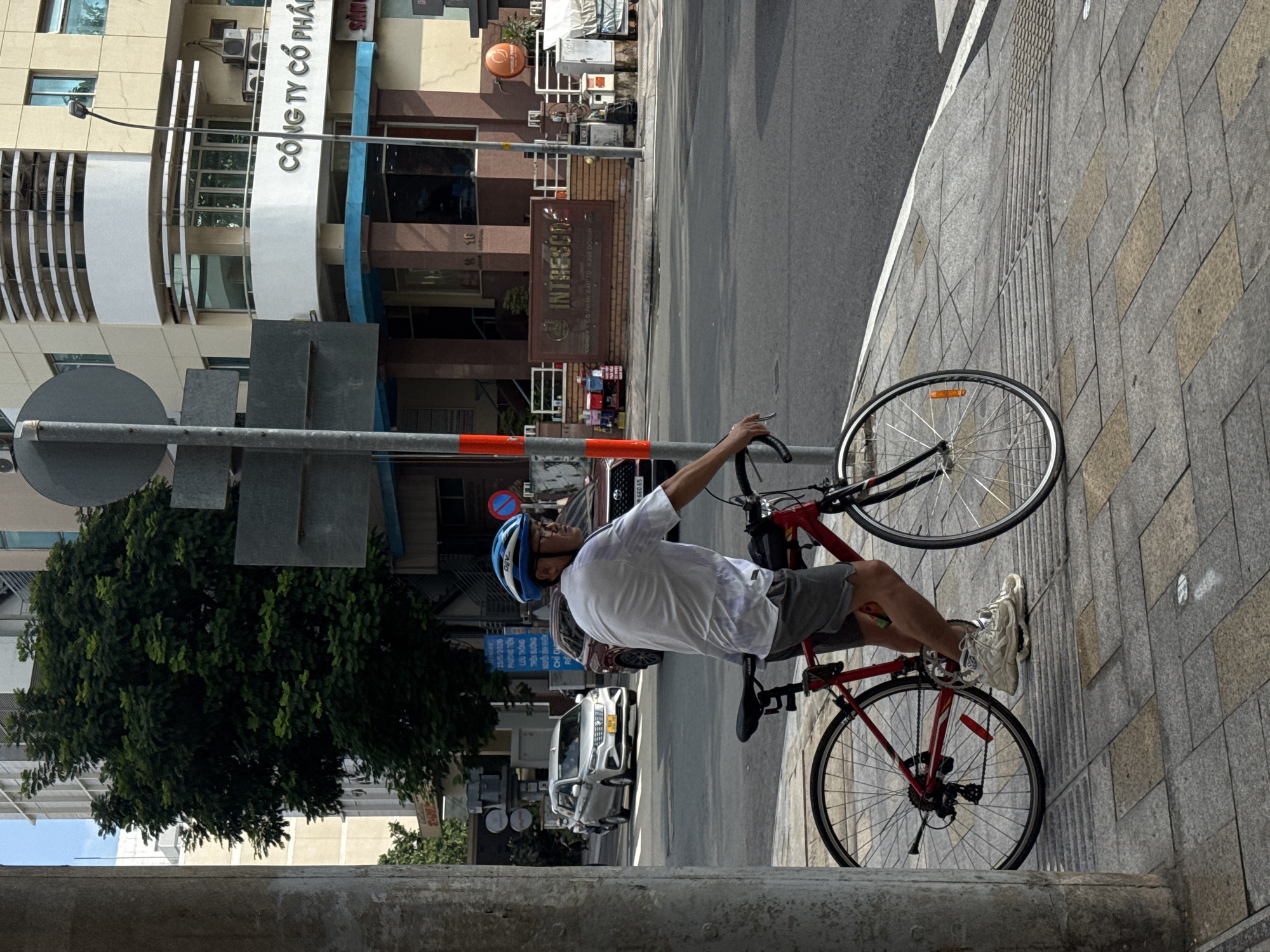 A man wearing a blue helmet and a white t-shirt rides a red bicycle on a sidewalk in a city. He is wearing grey shorts and white sneakers. The bicycle has a black seat and handlebars. In the background, there is a white car, a dark-colored car, and a building with "CÔNG TY CỔ PHẦN" written on it. The sidewalk is paved with gray tiles and there is a street with traffic in the background.
