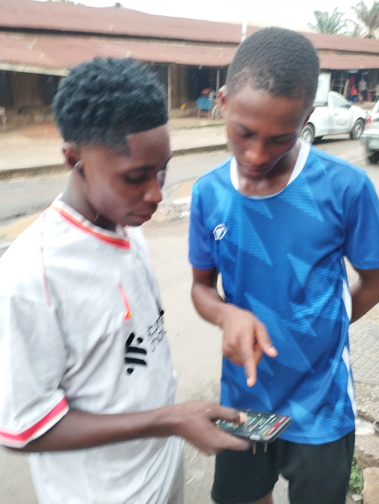 Two young men are standing on the side of a road, focused on a smartphone held by the individual on the left. The person holding the phone is wearing a white t-shirt with red and orange trim around the collar and sleeves. The other person is wearing a blue sports jersey. The location appears to be a street in Fegge, Nigeria, with buildings and vehicles visible in the background. The road surface looks wet. The smartphone screen is illuminated and shows various icons. One of the people is pointing at the screen.