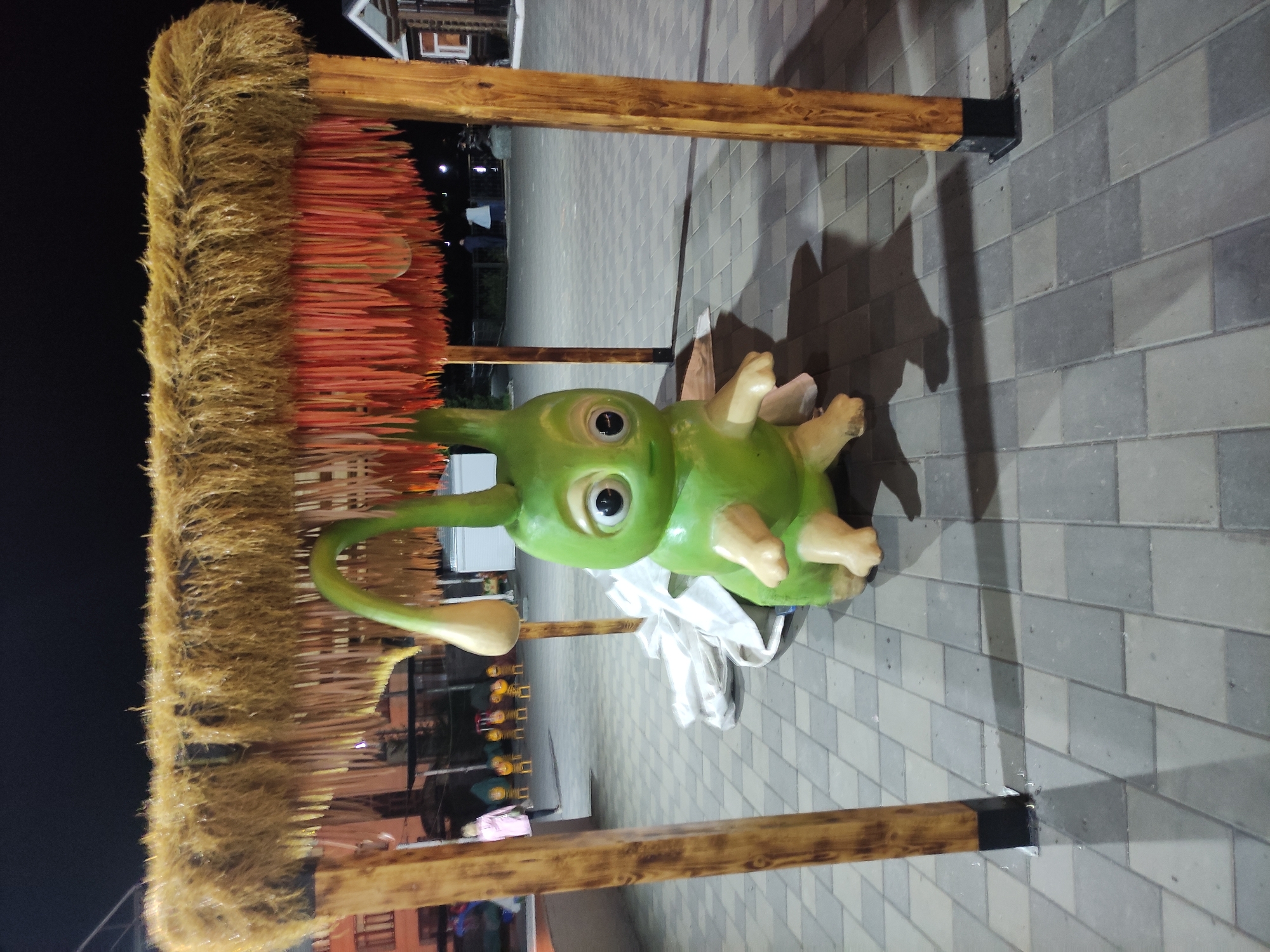 A ground-level, landscape-oriented image captures a whimsical scene on a paved surface. A cartoonish, green, alien-like figure with large, expressive eyes and pale limbs rests beneath a wooden frame structure. The frame supports a dense thatch or straw-like material, possibly forming a decorative awning. The paving stones are grey and laid in a rectangular pattern. The overall lighting suggests it is dusk or night. In the blurred background, hints of city activity, including lights and building structures, can be discerned. The scene suggests an outdoor public display, likely in a city like Pavlodar, Kazakhstan.