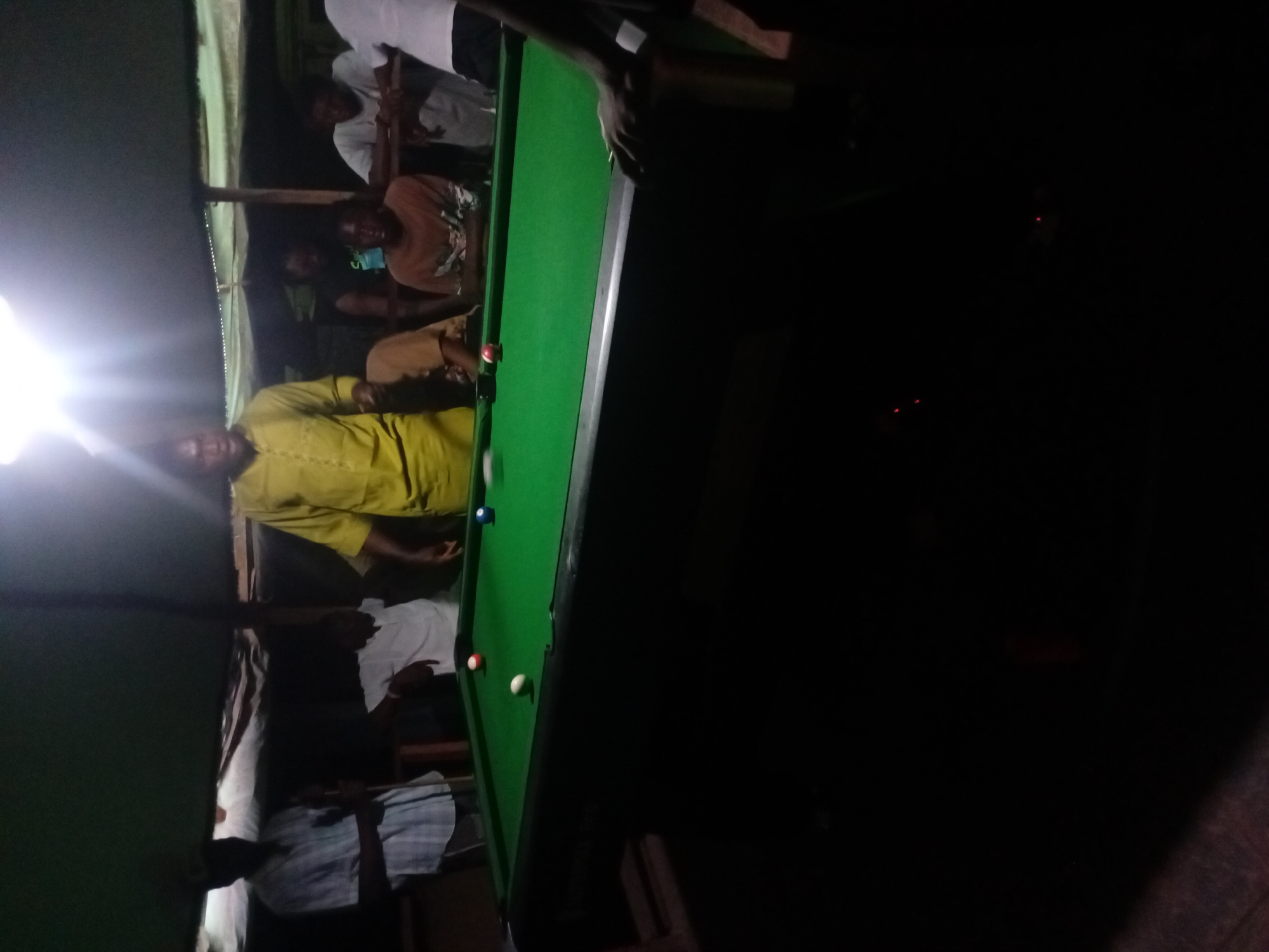 The image captures a dimly lit, informal gathering of several people around a pool table, suggesting a nighttime social activity. The photograph is rotated 90 degrees clockwise.
In the foreground and mid-ground on the left, a man wearing a bright yellow or lime green shirt stands next to the green-felt pool table, appearing to observe the game. Other individuals, mostly men, are partially visible in the background and foreground, their faces often obscured by shadows and poor lighting. One pair of hands is visible holding a pool cue stick over the table, indicating a player is either taking a shot or preparing to do so.
The central object is a green-felt pool or snooker table with visible wooden edges. Several pool balls are on the table, including a red ball, a white cue ball, and a blue ball. Much of the right side of the image is enveloped in darkness, with only faint glimmers of light visible in the deep background. A strong, bright artificial light source on the far left of the frame creates a significant lens flare and overexposure, contrasting sharply with the general dimness of the scene.
The activity taking place is a game of pool or snooker, with people gathered to play or watch. The setting appears to be an casual social venue, possibly an indoor game room or a covered outdoor space, characterized by wooden structures. The scene takes place at night, given the reliance on artificial lighting and the pervasive darkness. The atmosphere seems relaxed and social.
The event is set in Lokoja, Nigeria. There is no visible text in the image.