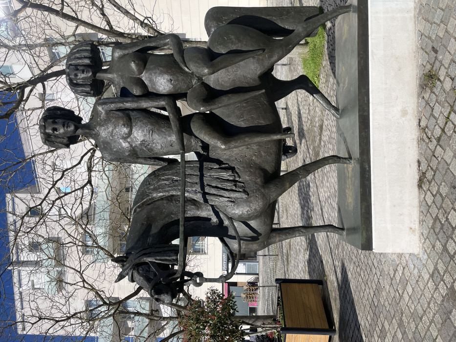 A bronze equestrian sculpture is depicted outdoors in Montigny-le-Bretonneux, France. The sculpture features a horse carrying two nude human figures. The male figure is positioned in front, sitting astride the horse, with stylized hair and lacking a right forearm. He holds reins with his left hand. The female figure is seated behind him in a side-saddle position, also nude, with a visibly pregnant abdomen and stylized hair. The bronze horse is bridled, with its right front leg raised, indicating a dynamic posture. The entire sculpture rests on a dark bronze plinth atop a rectangular, light-colored concrete pedestal. The ground is paved with small grey cobblestones. To the left, a wooden planter box contains a small shrub with reddish-green foliage. In the background, leafless deciduous trees partially obscure modern, multi-story buildings with light facades and balconies. The sky is clear blue, and strong sunlight illuminates the scene, casting distinct shadows.