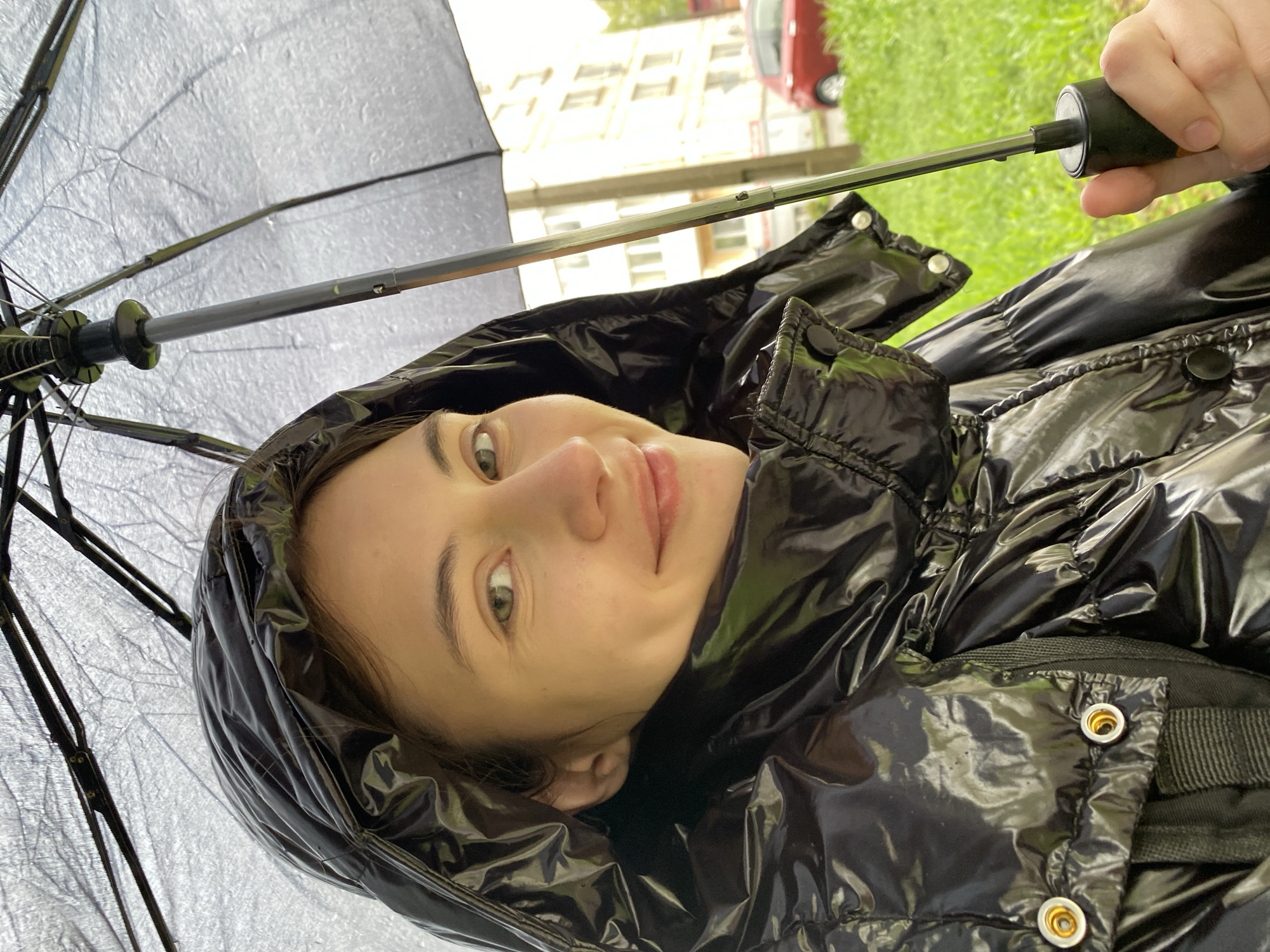 A woman in a black puffy jacket stands under a black umbrella, the edges of which frame the left side of the image. The woman is looking into the camera, smiling slightly. Behind her, out of focus, is a building with white walls and a few windows. A red car is visible partially obscured by the building. Grass is present in the background. Given the prompt's instruction to specify the city of Votkinsk, Russia, it is assumed the image was taken in this location.