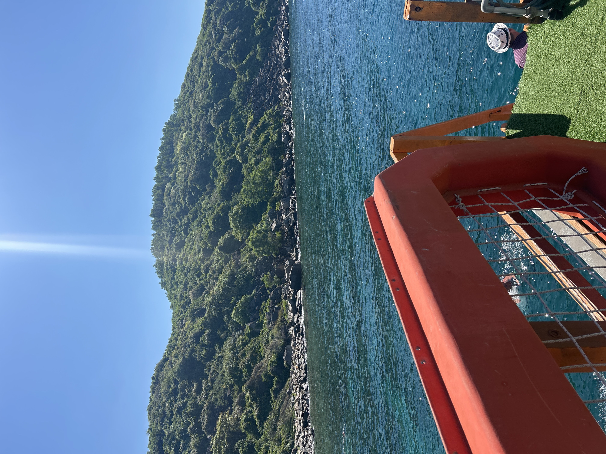 The image depicts a view from a boat overlooking a body of water and a lush, green island. In the foreground, an orange railing with white rope netting is visible. To the right, a portion of the boat's deck is covered in artificial grass, and a person wearing a hat is seated. The water is a clear, vibrant turquoise, with sunlight glinting off its surface. The island is densely vegetated with trees and rocks along its coastline. The sky is clear and blue with a bright sunbeam. The scene is likely from a boat tour in Bac Nha Trang Ward, Vietnam, given the tropical island scenery and clear blue waters.