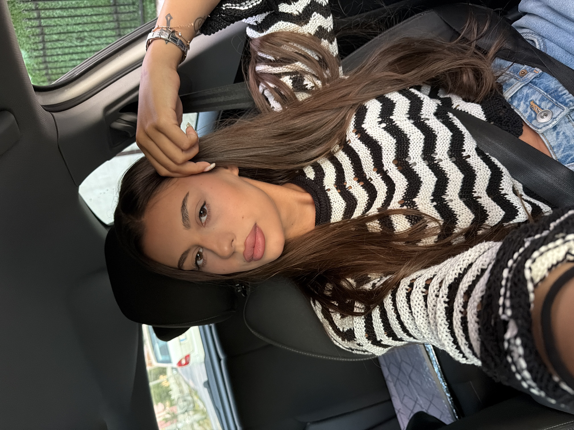 A young woman with long, dark brown hair and light skin is pictured inside a car, appearing to be in the driver's seat. She is wearing a black and white chevron-patterned knitted sweater and blue denim jeans. Her right hand is resting on her forehead, and she is looking towards the camera. The interior of the car is visible, including a black leather seat and a seatbelt. Outside the car window, a street scene with buildings and a white vehicle can be observed. There is no specific visual information in the image to indicate the location is Karnobat, Bulgaria.