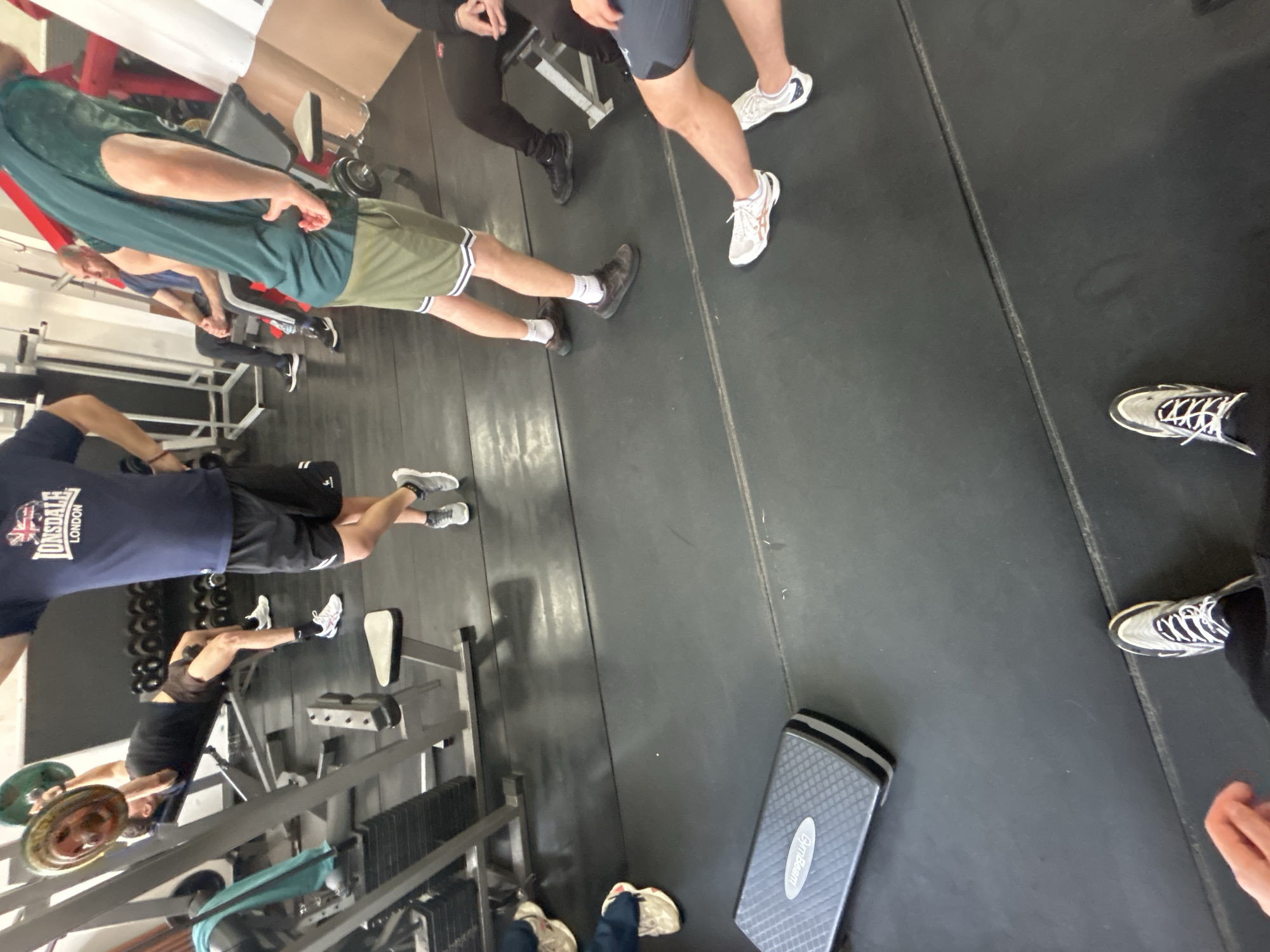 The image shows the interior of a gym in Zlatograd, Bulgaria. Several people are present, some standing and others using gym equipment. A man is lifting weights on a bench. Another person is wearing a Lonsdale t-shirt. There is a step platform on the floor. The flooring appears to be rubberized. Weight plates are visible on a rack. Gym equipment, including benches and weight machines, are scattered throughout the space.