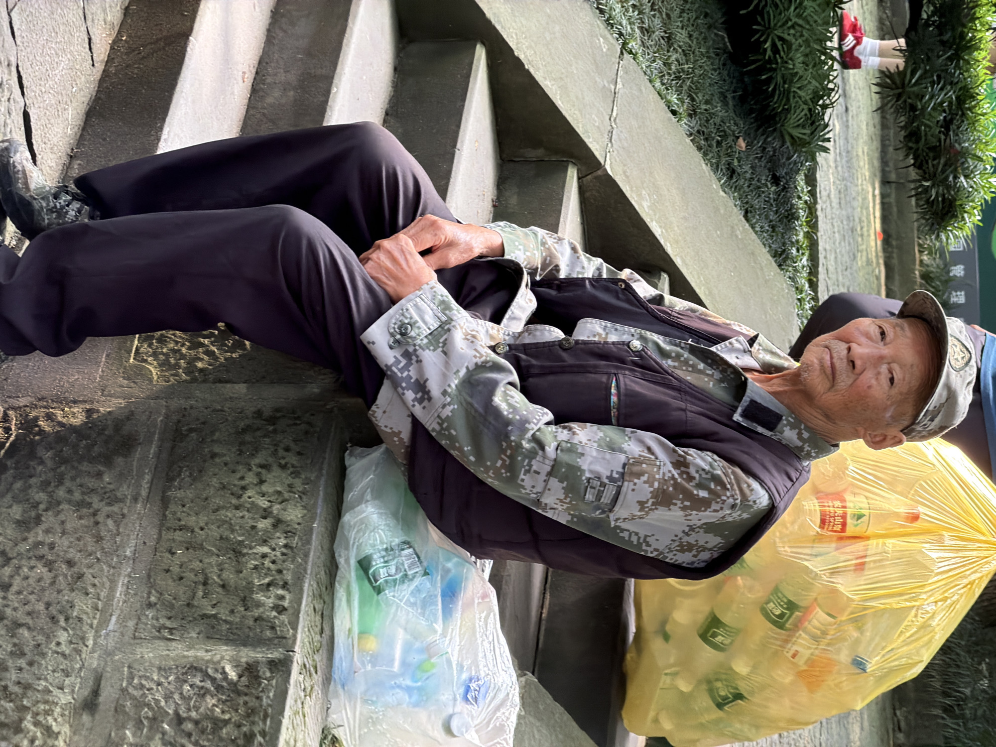 An elderly East Asian man is seated on a stone ledge in an outdoor park setting in Yuzhong District, China. He wears a camouflage long-sleeved shirt, a dark vest, and dark pants. A light-colored cap with an emblem sits on his head. His hands are clasped in his lap. To his right, a large, translucent yellow plastic bag is filled with plastic bottles. In front of him and to his right, a clear plastic bag also contains bottles and other items. Behind him, steps lead up to a grassy area with bushes. The man is looking directly at the camera with a neutral expression.