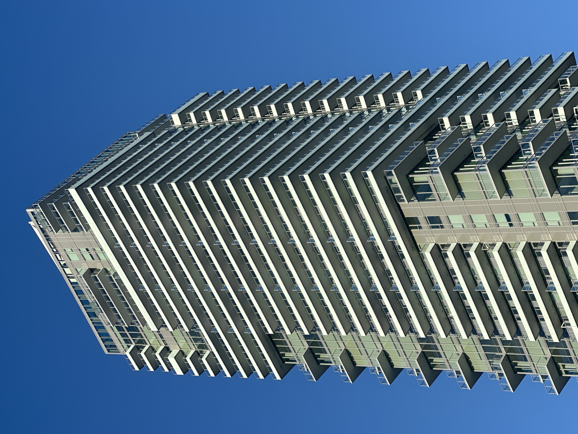 The image shows a modern high-rise residential building against a clear blue sky. The building features a prominent, repeating design of white balconies with glass railings that wrap around multiple floors. A section of the building on the left side has a glass facade. The building appears to be located in an urban setting, as suggested by its architecture. Based on public records and architectural styles, this building is likely the "Esprit II" condominium tower located at 3099 Glen Drive in Coquitlam, British Columbia, Canada. There are no people visible in the image, and the scene is static. The environment is characterized by bright daylight and a cloudless sky.