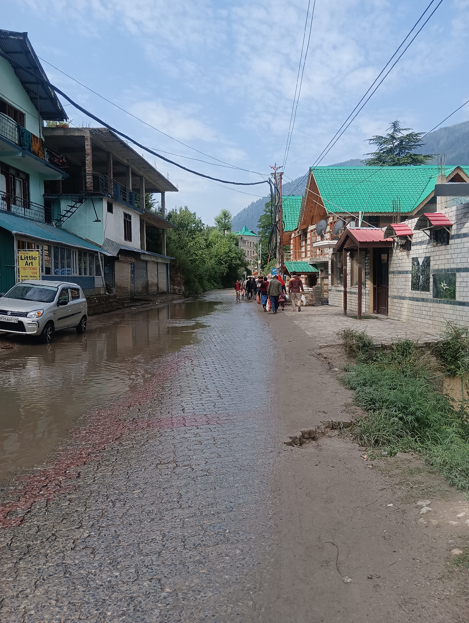 In Mujuri, India, a street scene unfolds under a cloudy sky. The road is partially submerged in water, reflecting the surroundings. On the left, a light-colored car is parked near a building with blue and white siding, featuring multiple balconies. A sign for "Little Art Traditional Food" is visible on this building. Adjacent to it are shops with closed metal shutters. Further down the street, a group of people, some carrying bags, walk away from the camera. The right side of the street features buildings with green tiled roofs, wooden accents, and brickwork. Lush green foliage grows along the side of the road. The overall impression is of a wet, possibly recently rained-upon, urban environment.