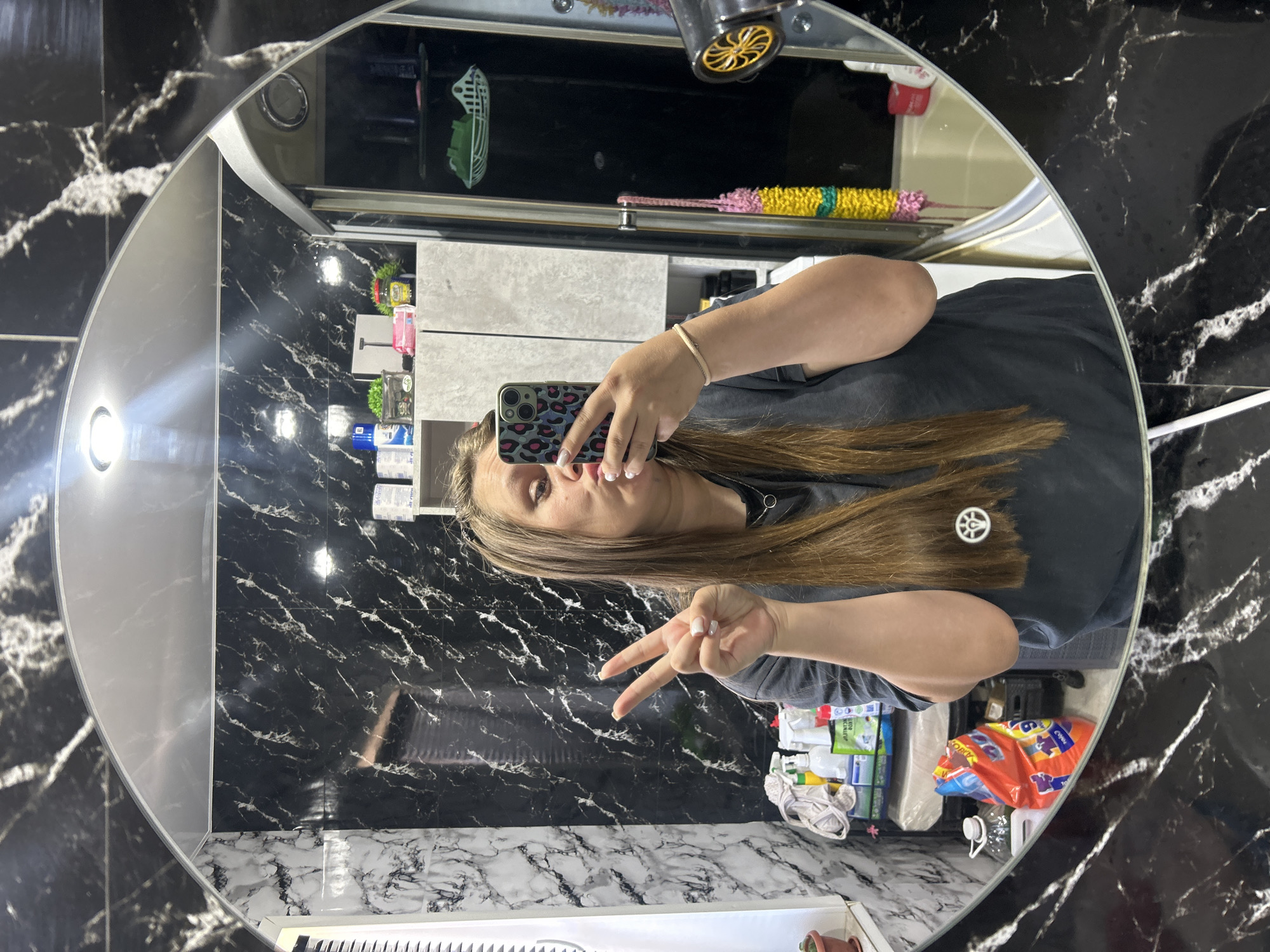 The image shows a young woman taking a mirror selfie in a bathroom. The bathroom features black and white marble-patterned tiles on the walls and a silver-colored arch above the mirror. The woman is wearing a dark gray t-shirt and has long, brown hair. She is making a peace sign with her left hand and holding a smartphone with a leopard-print case in her right hand. In the background, reflected in the mirror, are various bathroom items including cleaning supplies, toiletries, a shower curtain, and a bathtub. The overall setting appears to be a domestic bathroom, possibly in a residential building. The location of Ольховка, Russia is not visually determinable from the image itself.