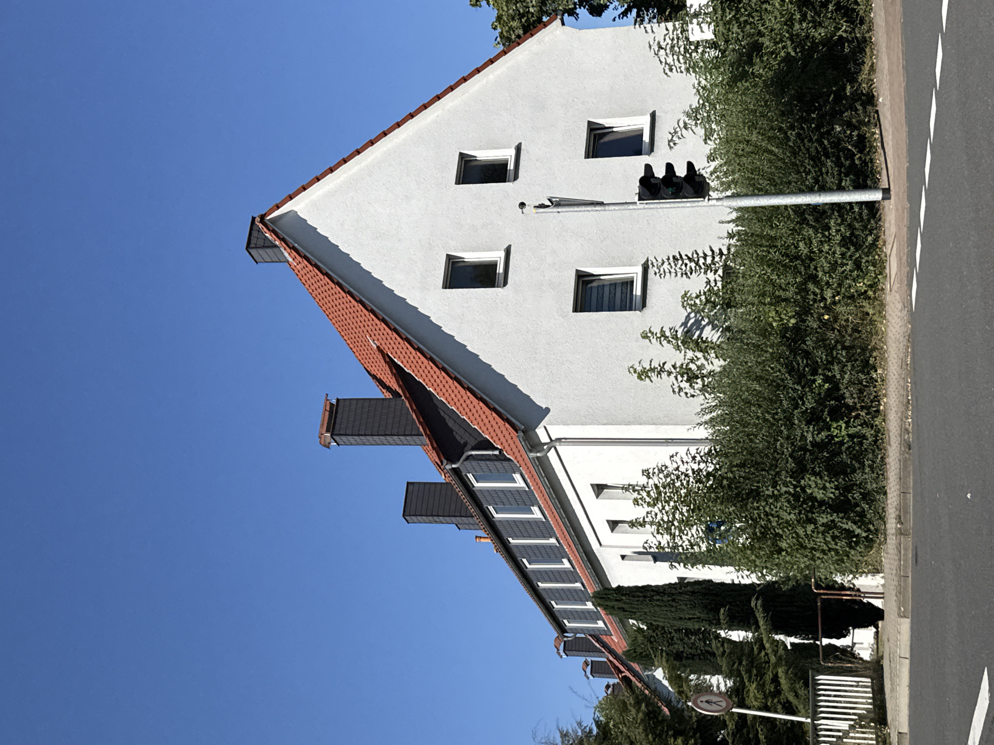 The image shows a residential building in Herzberg, Germany, under a clear blue sky. The building has a white facade and a red tiled roof. There are several windows visible, some with dark frames and others with white frames. Chimneys with dark slate cladding protrude from the roof. In front of the building, there is a line of lush green bushes and trees. A traffic light is positioned on the sidewalk, with its lights illuminated green, yellow, and red, indicating a functioning traffic signal. A white metal fence and a pedestrian crossing sign are also visible along the sidewalk. The street in the foreground is asphalt, with white dashed lines marking the lanes. The scene appears to be a typical urban or suburban streetscape on a sunny day.