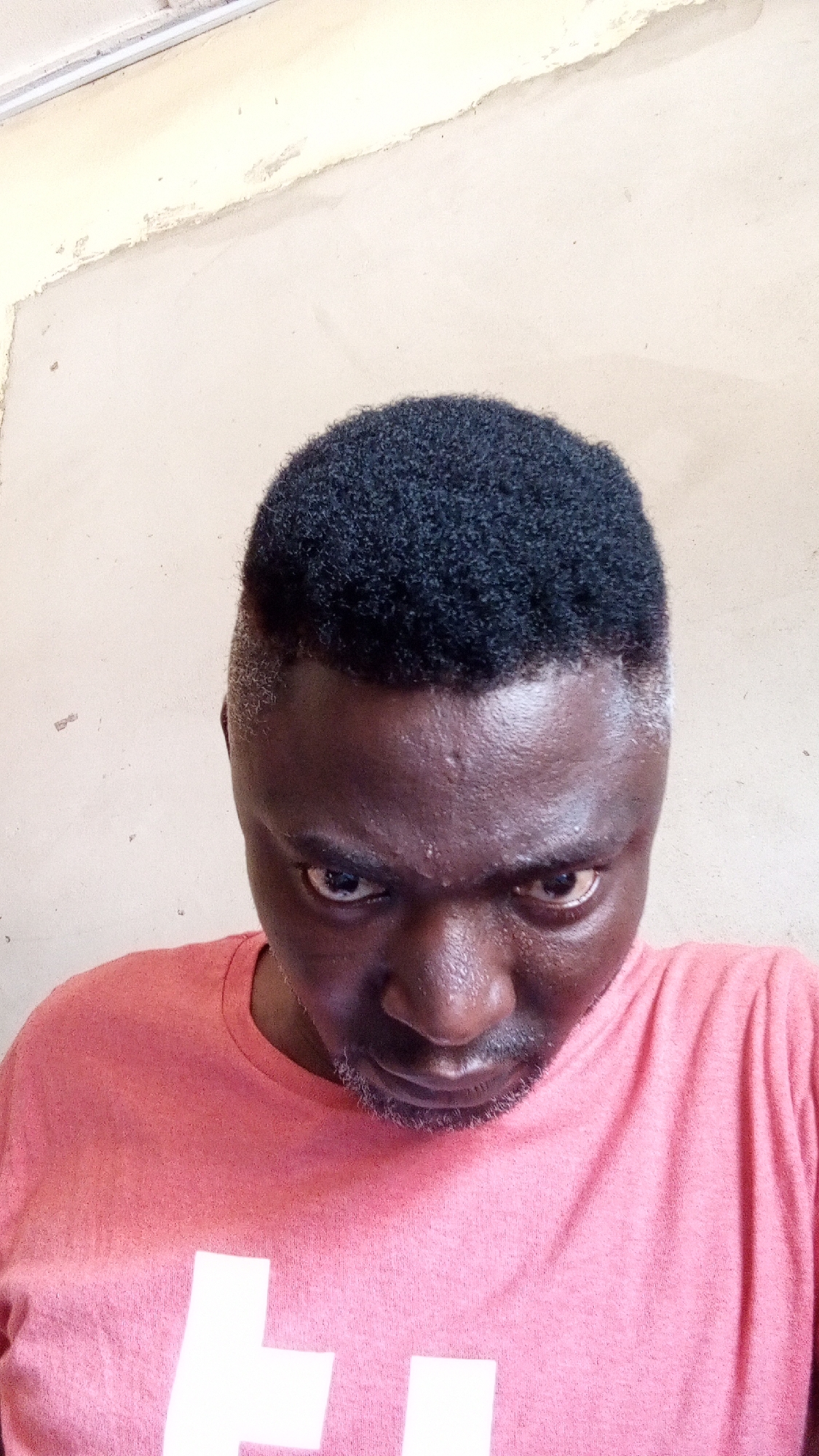 The image presents a close-up, high-angle portrait of an adult male. He has dark skin, short, dense black curly hair on top, and closely shaved sides with a distinctive fade. A short beard or stubble covers his chin and jawline. His eyes are wide open, and his brow is furrowed, conveying an intense, serious, or possibly determined gaze directed straight at the viewer. He is wearing a coral or salmon pink crew-neck t-shirt, which has a partial white, block-letter graphic visible towards the bottom of the frame, resembling a capital 'H' or similar shape, possibly part of a logo or text. The background is a plain, light-beige or off-white wall showing some patches of peeling paint and general wear, suggesting an aged indoor or sheltered outdoor setting. The lighting is bright and diffused, indicative of daytime. The man appears to be posing for a photograph, likely a selfie, given the perspective.