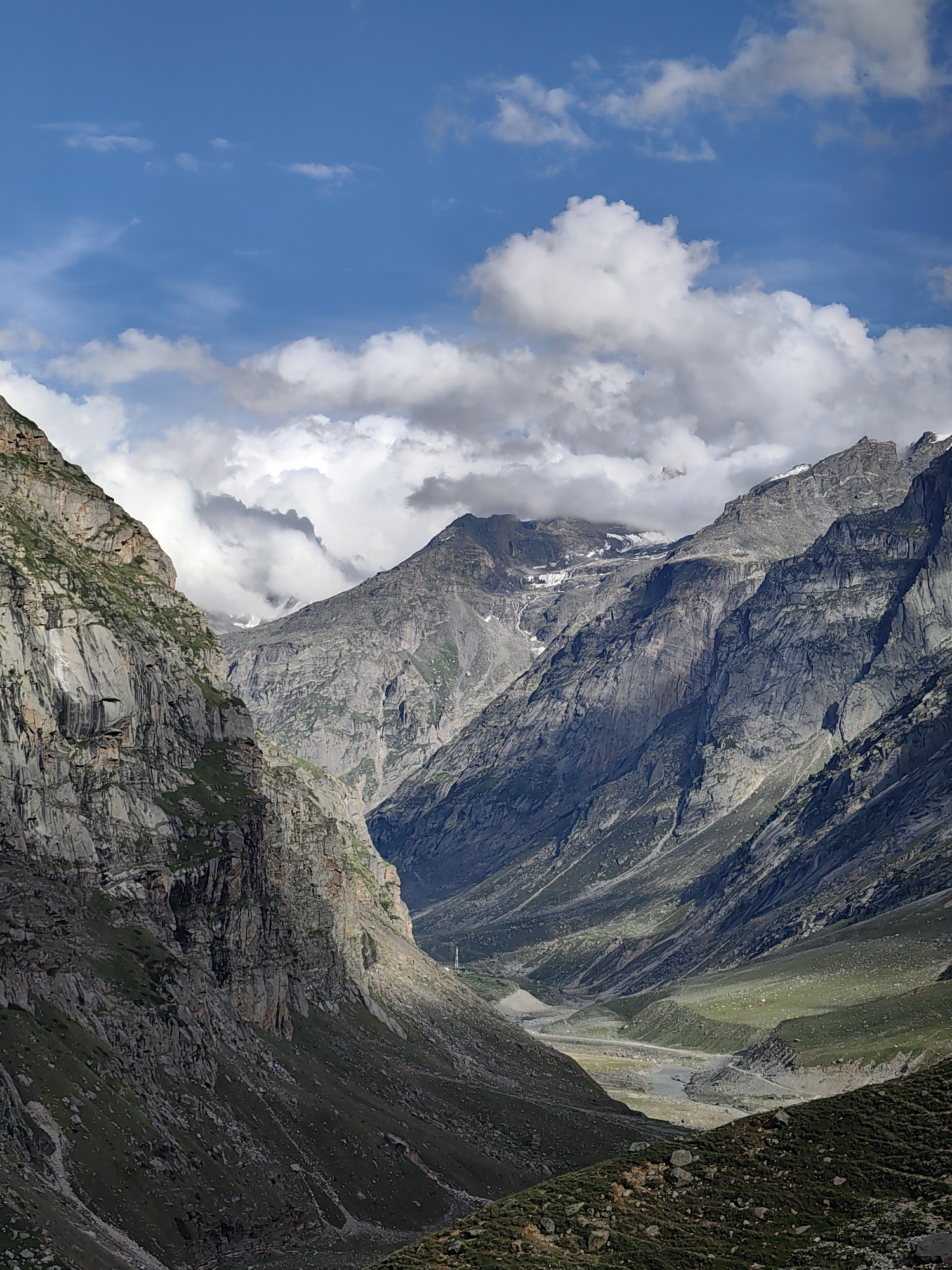 The image depicts a majestic mountain landscape, likely in the vicinity of Bashisht, India, given the characteristic terrain. The scene is dominated by steep, rocky mountainsides, some covered with patches of sparse green vegetation. A valley carved by a river or stream stretches into the distance, with its bed appearing light-colored and rocky. The sky above is a vibrant blue, punctuated by dramatic white and gray clouds that cast shadows on the mountains, creating a play of light and dark across the rugged terrain. The overall impression is one of immense natural beauty and geological grandeur.