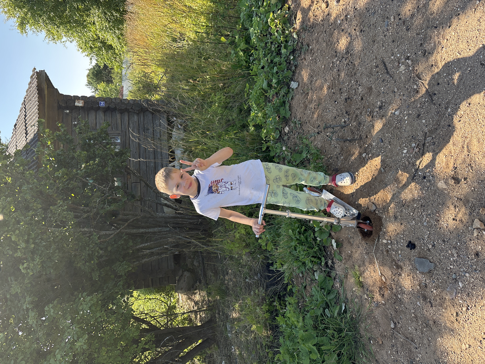 A young boy stands on a dirt path in front of a rustic wooden house in the village of Dunilovo, Russia. He is wearing a white t-shirt and light green pants with a cartoon pattern. He is holding a silver scooter and making a peace sign with his right hand. The house is made of dark logs and has a corrugated roof. The area is overgrown with tall grass and trees. The sun is shining, casting long shadows.