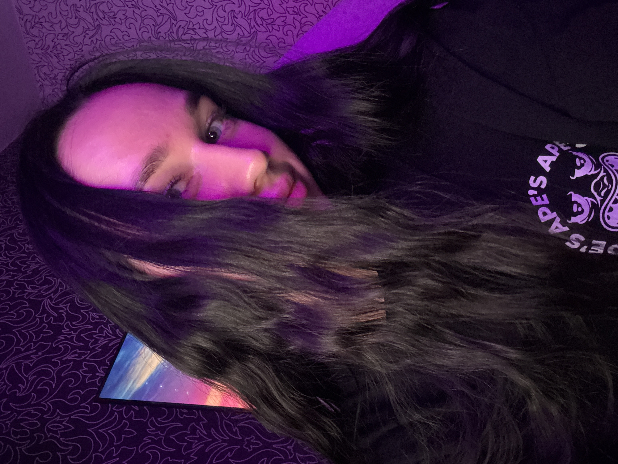 The media file depicts a close-up, low-angle shot of a person lying down, with their head tilted and partially covered by dark hair. The scene is dimly lit, with a strong purple light source casting a vibrant glow on the person's face and hair. The background features a patterned fabric, likely a bedsheet or wallpaper, in a dark purple hue. To the left of the frame, a portion of a device screen is visible, displaying a colorful image of a sky with clouds and streaks of light. The person appears to be looking towards the viewer, with one eye visible through their hair. Their expression is somewhat neutral, but the lighting creates an atmospheric and perhaps intimate mood.
On the person's clothing, a circular graphic is visible with text. The visible text reads "APE'S AP" and "ES AP E'S A". The overall aesthetic is somewhat moody and artistic, possibly taken indoors at night given the artificial lighting.
No specific weather or explicit Novosibirsk location cues are present beyond the provided context. The activity appears to be a person relaxing or posing for a photograph, possibly while looking at or interacting with the device.