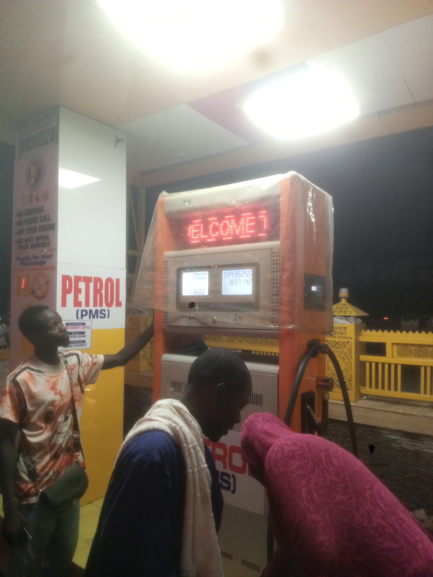 The image shows a petrol (PMS) station in Gwarinpa, Nigeria, at night. A fuel pump with "WELCOME!" in red LED display shows the current fill: 20000 and the total cost: 13680. A man wearing a printed shirt and a crossbody bag is standing to the left of the pump. In front of the pump, another man with a white towel around his neck is wearing a blue shirt and a person wearing a pink hijab is facing the pump. A sign next to the pump displays rules and a contact number. The station is identified by a sign, featuring the word "PIRE", "ERGY" and "FILLING STATION"