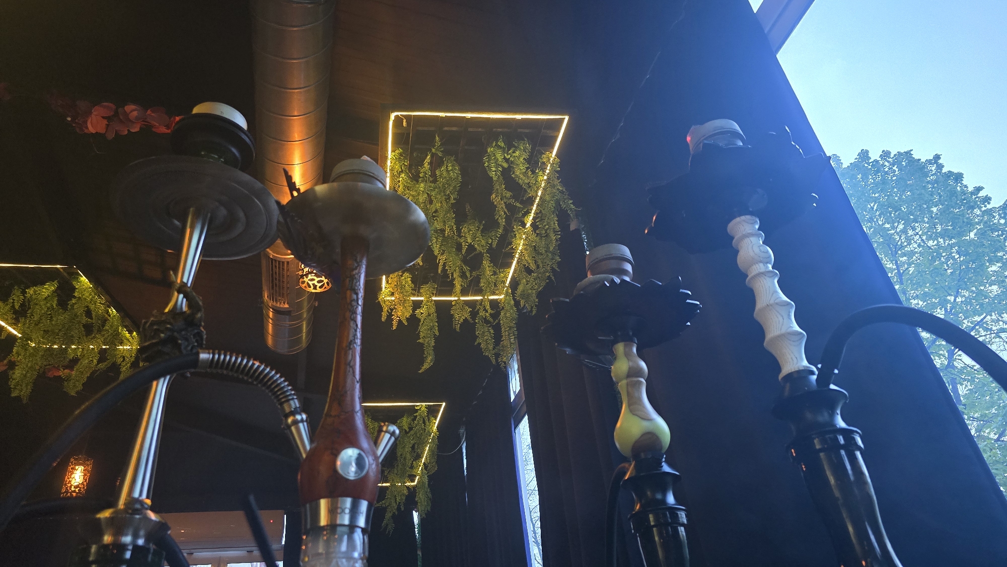 This media file presents an atmospheric interior view of what appears to be a shisha lounge or cafe, possibly in Sofia, Bulgaria. The image is taken from a low angle, looking upwards, emphasizing the height and design of multiple hookahs.
In the foreground and mid-ground, several hookahs stand prominently. They feature varied designs: one has a metallic, industrial appearance with a ribbed hose, while another sports a reddish-brown wooden stem with a silver metallic base, where the word "VOODOO" is faintly visible. A third hookah is predominantly black with a distinct white, ridged section on its stem. The hookahs appear ready for use, with their bowls visible at the top.
The setting combines industrial and natural design elements. Above, exposed silver ventilation ducts run across the ceiling, illuminated by a warm, orange light. Geometric frames of bright neon yellow light are suspended from the dark ceiling, enclosing vibrant green artificial hanging plants, creating an artistic and modern decorative effect. To the right, a large window reveals a bright, clear blue sky and the green foliage of a tree, indicating it is daytime or early evening outside. Dark curtains or walls frame this outdoor view, contrasting with the interior's artificial lighting. A small, decorative light fixture with a geometric pattern is also visible in the distance.
No people or specific activities are shown, allowing the focus to remain on the establishment's distinct decor and its array of hookahs, suggesting a relaxed and trendy environment for leisure.