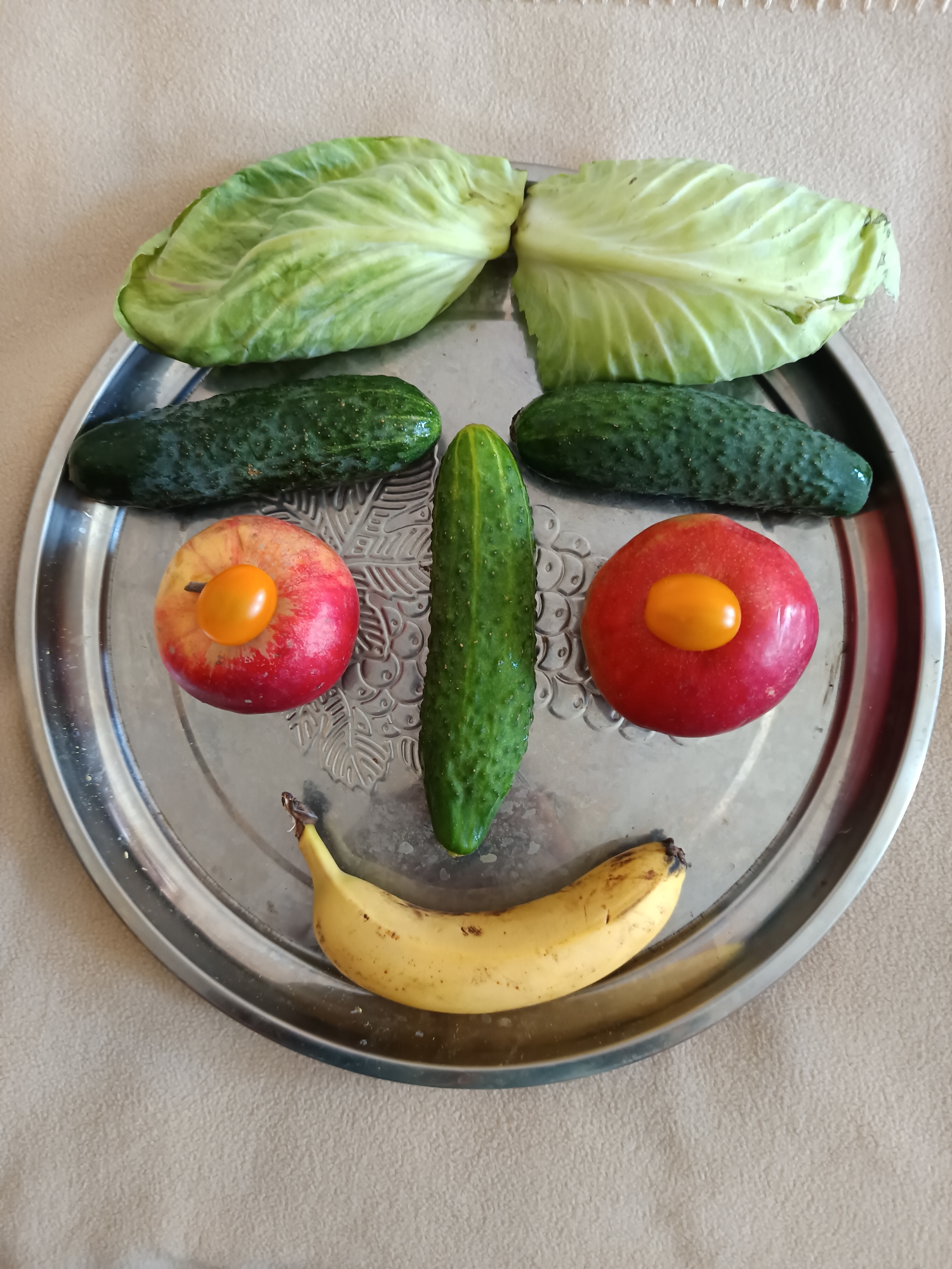 A face made of fruits and vegetables is arranged on a metallic plate. The face has two cabbage leaves for eyebrows, two cucumbers for eyes, two apples with yellow cherry tomatoes for cheeks, a cucumber for a nose, and a banana for a mouth. The plate is placed on a light brown, textured surface. There is no indication of the location in Tyshky, Ukraine.