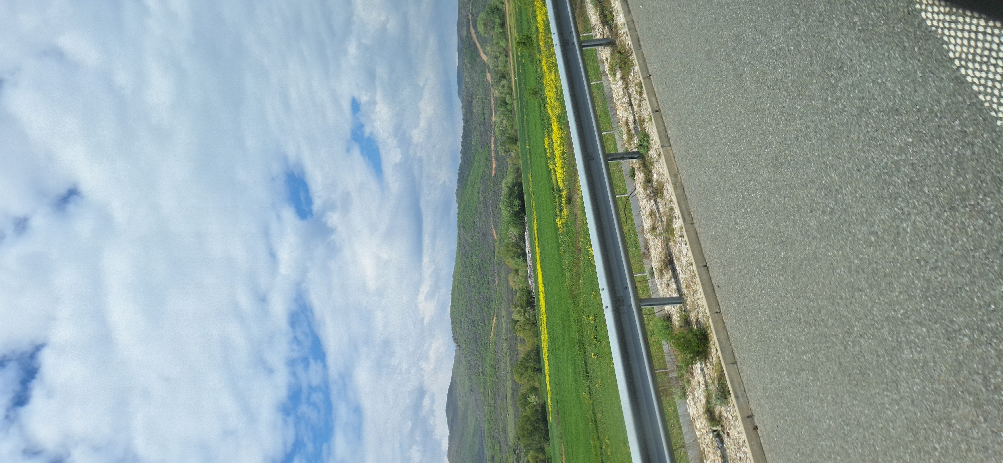 The image shows a roadside scene near Valkovo, Bulgaria. A gray asphalt road occupies the right side of the frame, with a metal guardrail separating it from a green field dotted with yellow flowers. Beyond the field is a hillside covered in green vegetation. The sky above is blue with scattered white clouds.