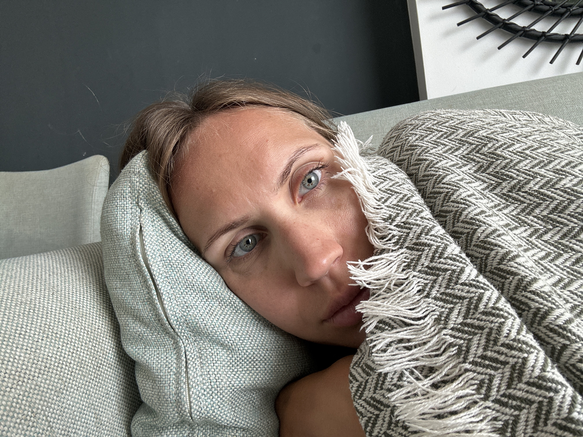 A woman is reclining on a light blue upholstered sofa. She is resting her head on a light blue pillow and partially covered with a gray and white herringbone patterned blanket with white fringe. Part of her face is obscured by the blanket. The background includes a dark gray wall and part of a white wall with a black circular decorative object. The location in Fethiye, Turkey, cannot be determined based on the visual information.