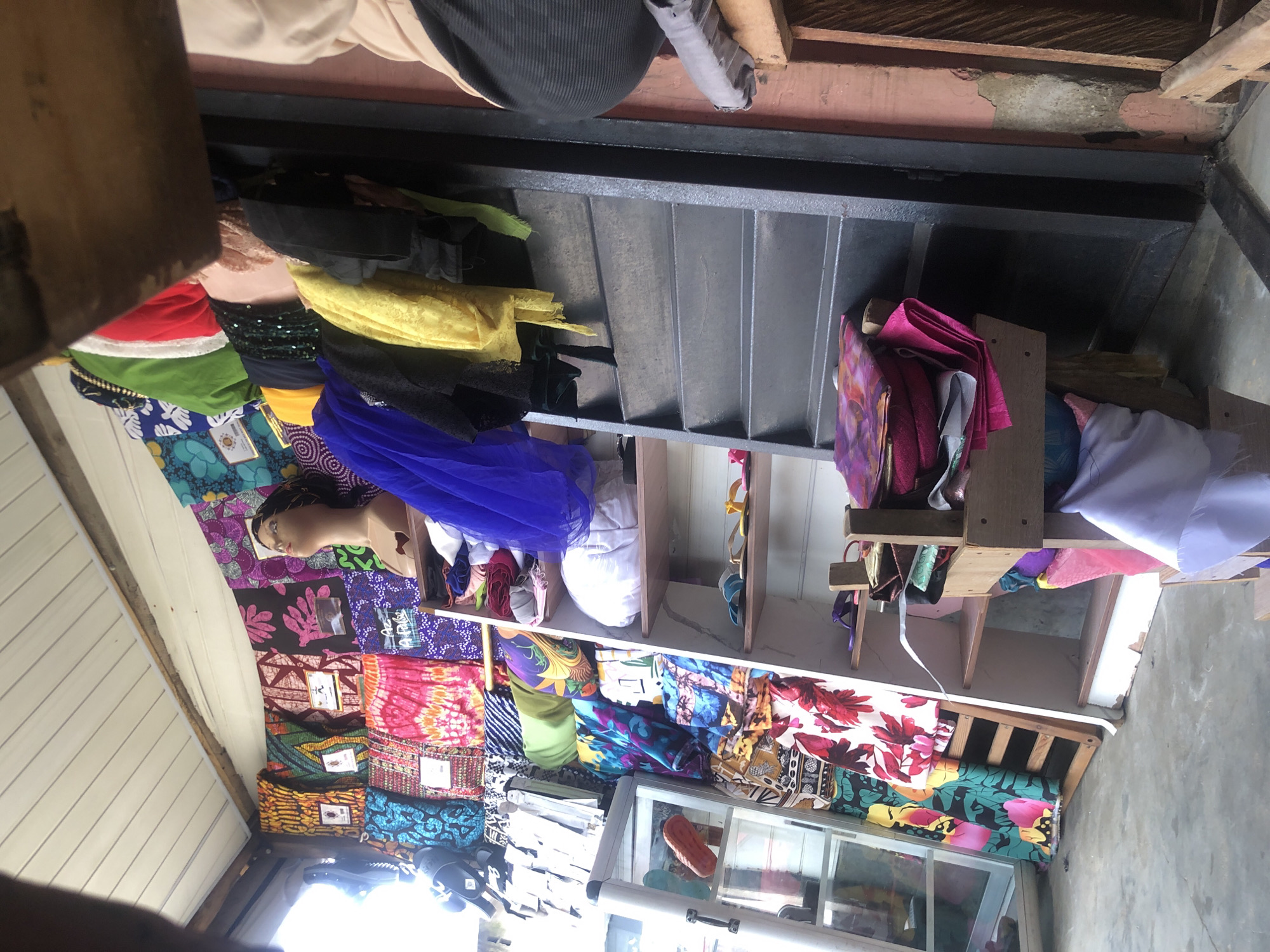 The image depicts a cluttered shop interior, likely in Ado, Nigeria, filled with a vibrant assortment of fabrics and clothing. In the foreground, a person's legs and torso are visible, wearing dark, striped trousers and a light-colored garment. They are positioned to the right of the frame, partially obscuring the scene. The shop features numerous shelves laden with colorful textiles, including patterned fabrics, dresses, and what appear to be scarves or wraps. A mannequin head, with dark hair, is placed prominently among the displayed items. Various racks and display shelves are packed with merchandise, creating a dense visual field. The lighting suggests an indoor setting, possibly illuminated by natural light filtering in from an unseen window or opening. The overall impression is of a busy, well-stocked marketplace stall specializing in fabrics.