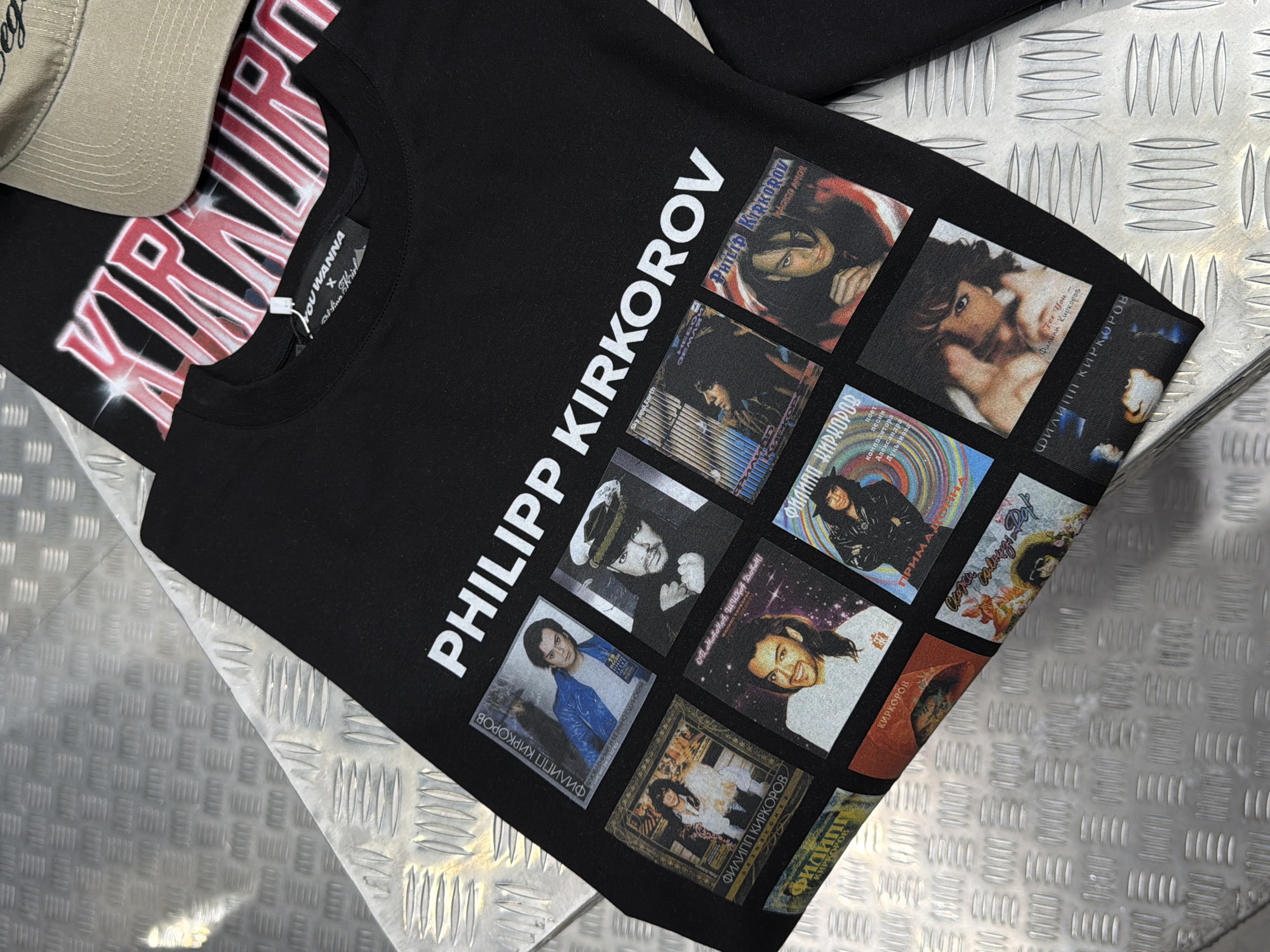 This image shows a black t-shirt with a graphic print of the Russian singer Philipp Kirkorov. The print features multiple square images of Kirkorov in various outfits and poses, some resembling album covers. The text "PHILIPP KIRKOROV" is printed in large white letters diagonally across the shirt. Smaller text, "YOU WANNA" and "Sinn Kirl", along with Russian text that translates to "For You Philipp Kirkorov", "Prima Donna", and other song titles, are visible on the individual images. A tan baseball cap with embroidered text is partially visible at the top left. The t-shirt is laid flat on a textured metallic surface, possibly a table or floor, with a checkered pattern, common in industrial or workshop settings. The lighting suggests an indoor environment. The overall presentation is that of merchandise for sale or display.