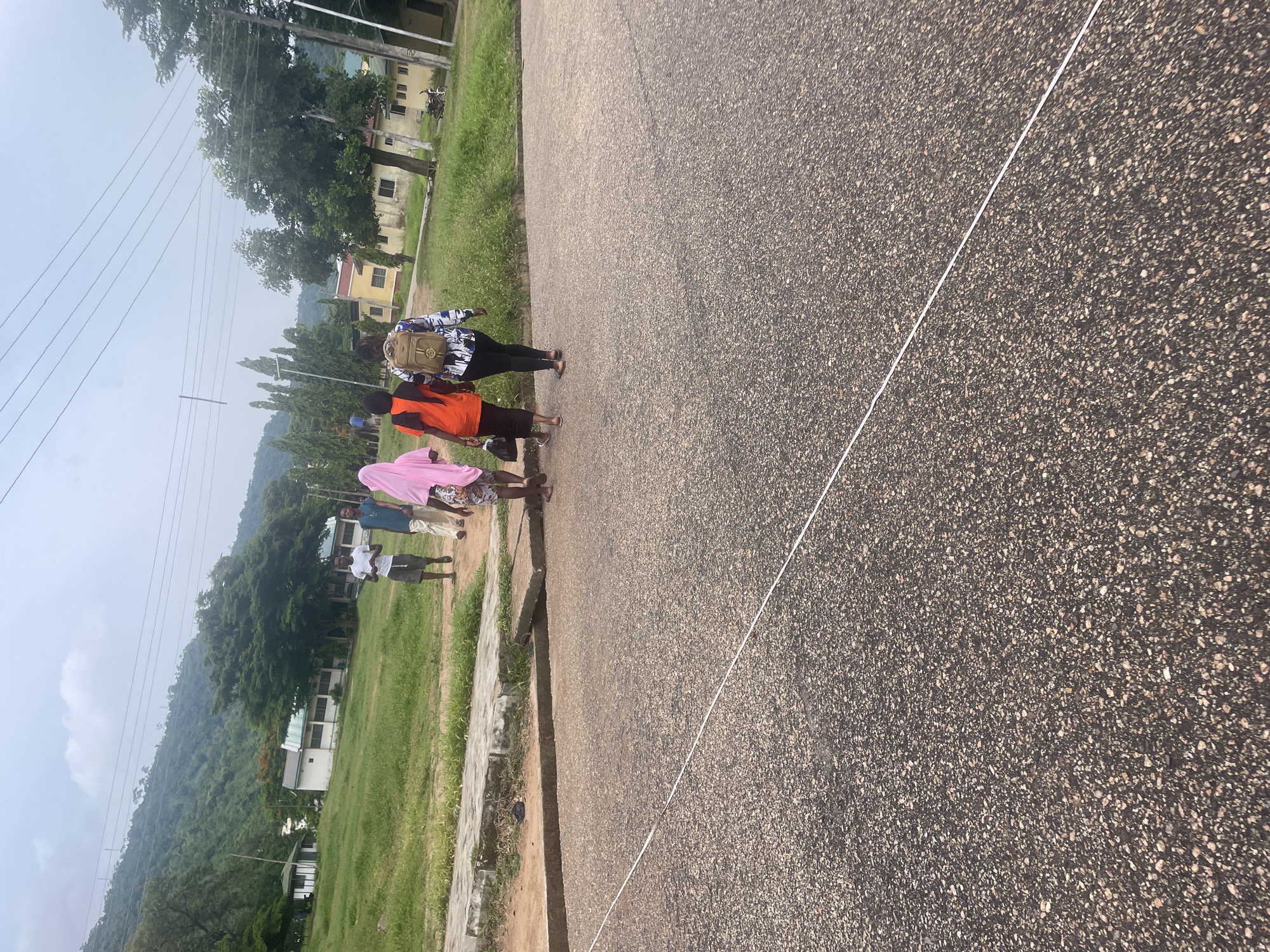 The image depicts a group of four individuals walking on a paved road in Lokoja, Nigeria. The scene is set against a backdrop of lush green hills and vegetation, with several buildings visible in the distance. The sky is overcast, suggesting a cloudy day.
In the foreground, the road surface is rough asphalt with a thin white line running diagonally across it. To the left of the road, there is a concrete sidewalk with some overgrown grass and a raised concrete structure.
The group of four comprises two men and two women, all walking away from the camera. The two men are at the front, one wearing a white t-shirt and shorts, and the other in a blue t-shirt and light-colored trousers. The two women are behind them. One woman is wearing an orange top and black trousers, and the other is wearing a patterned blue and white shirt and black trousers, with a beige backpack slung over her shoulders. The woman on the far left is wearing a pink headscarf and a patterned pink and white garment.
In the background, there are several buildings, appearing to be part of an institutional or residential complex. Trees and other vegetation are scattered throughout the landscape, contributing to the verdant appearance. Power lines are visible stretching across the upper portion of the image.