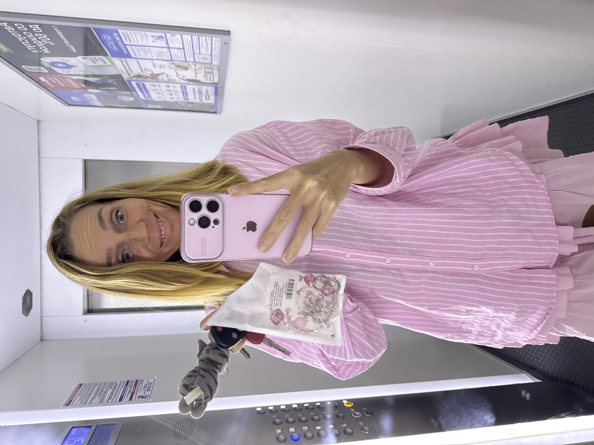 A woman stands in an elevator, holding a pink iPhone and a bag of jewelry. She is wearing a pink and white striped button-down shirt and a layered pink dress. A keychain with a cat charm is attached to her left hand. The elevator has a panel of buttons on the left wall. A framed poster is visible on the right wall, with Cyrillic text. The woman is smiling at the camera.
