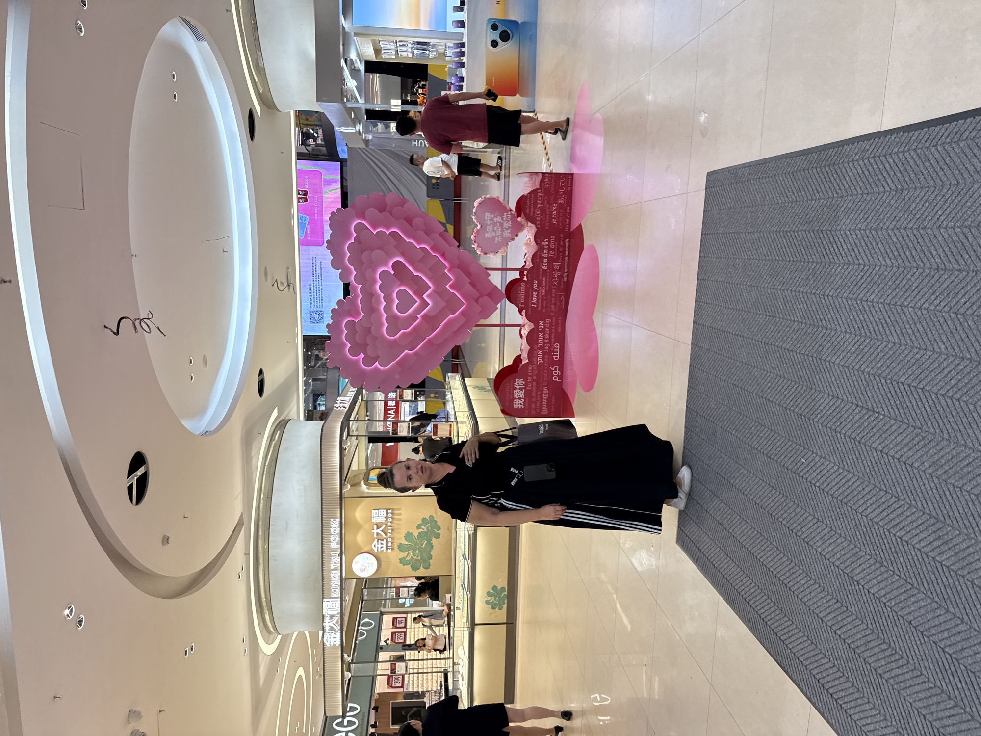 The image shows the interior of a shopping mall in the Tianhe District of China. A woman in a black dress with white stripes on the sleeves and a purse over her shoulder is standing to the right of a pink heart-shaped balloon display with "I love you" written in different languages. The text on the pink sign appears to be advertising the theme of the balloon display. Behind the woman is a storefront with the name "KING TAI FOOK" in Chinese characters. There are people shopping inside the store. The ceiling of the mall is high with oval-shaped light fixtures. A gray mat covers a portion of the floor in the foreground. In the background, there are other stores and people walking through the mall.