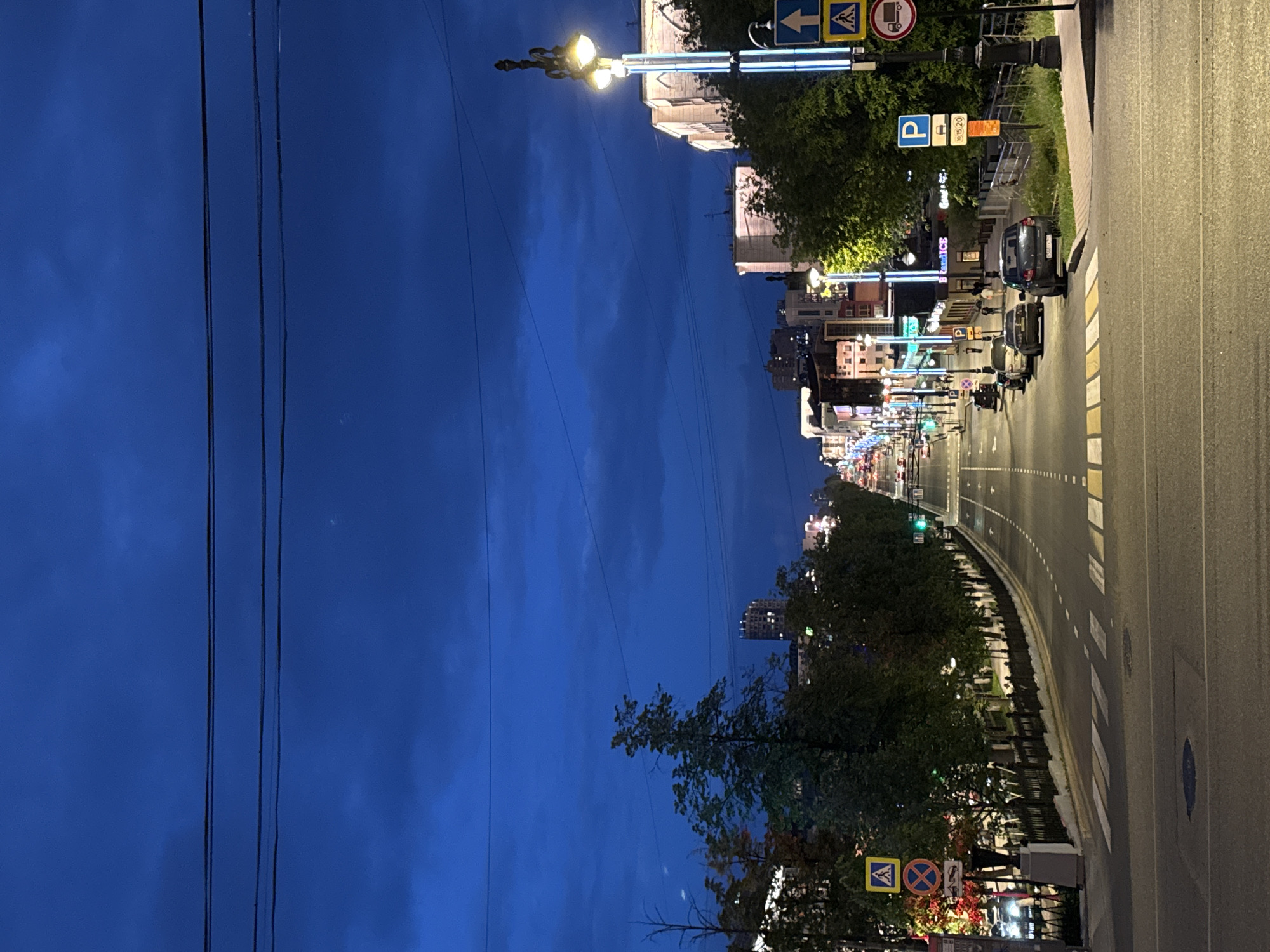 The image depicts a street scene in the city of Perm, Russia, at dusk or early night. The sky is a deep, dark blue with some clouds visible. Overhead, multiple power lines crisscross the frame. The street is a multi-lane road with traffic lights and lane markings. There are cars parked along the side of the road and some driving on it. Pedestrian crossings are marked with white stripes on the asphalt.
Along both sides of the street, there are buildings with illuminated windows and signs, indicating businesses or residences. Trees line the sidewalks, some with autumn foliage. Streetlights, some with ornate designs and others with modern vertical lighting, illuminate the street. Several traffic signs are visible, including a pedestrian crossing sign, a parking sign, and a truck restriction sign.
In the distance, the street continues and appears to be lined with more buildings and lights, suggesting an urban environment. The overall impression is one of a city street transitioning from daylight to night, with the artificial lights of buildings and streetlamps beginning to dominate the scene.