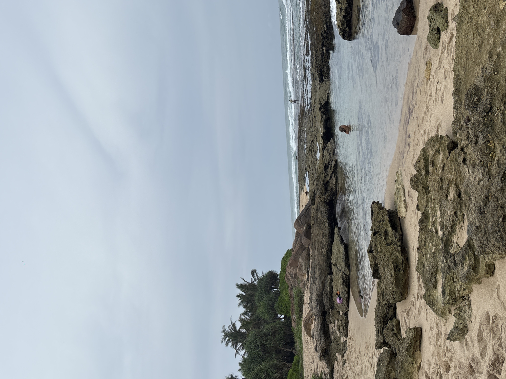 The image depicts a coastal scene in Thalaramba, Sri Lanka. The sky is overcast. The foreground features a sandy beach with large, dark, weathered rocks. A pool of water is enclosed by these rocks, in which a person is bathing. Further out, the ocean's waves break against the shoreline. A second individual is standing on the shoreline. Palm trees grow on the land adjacent to the beach.