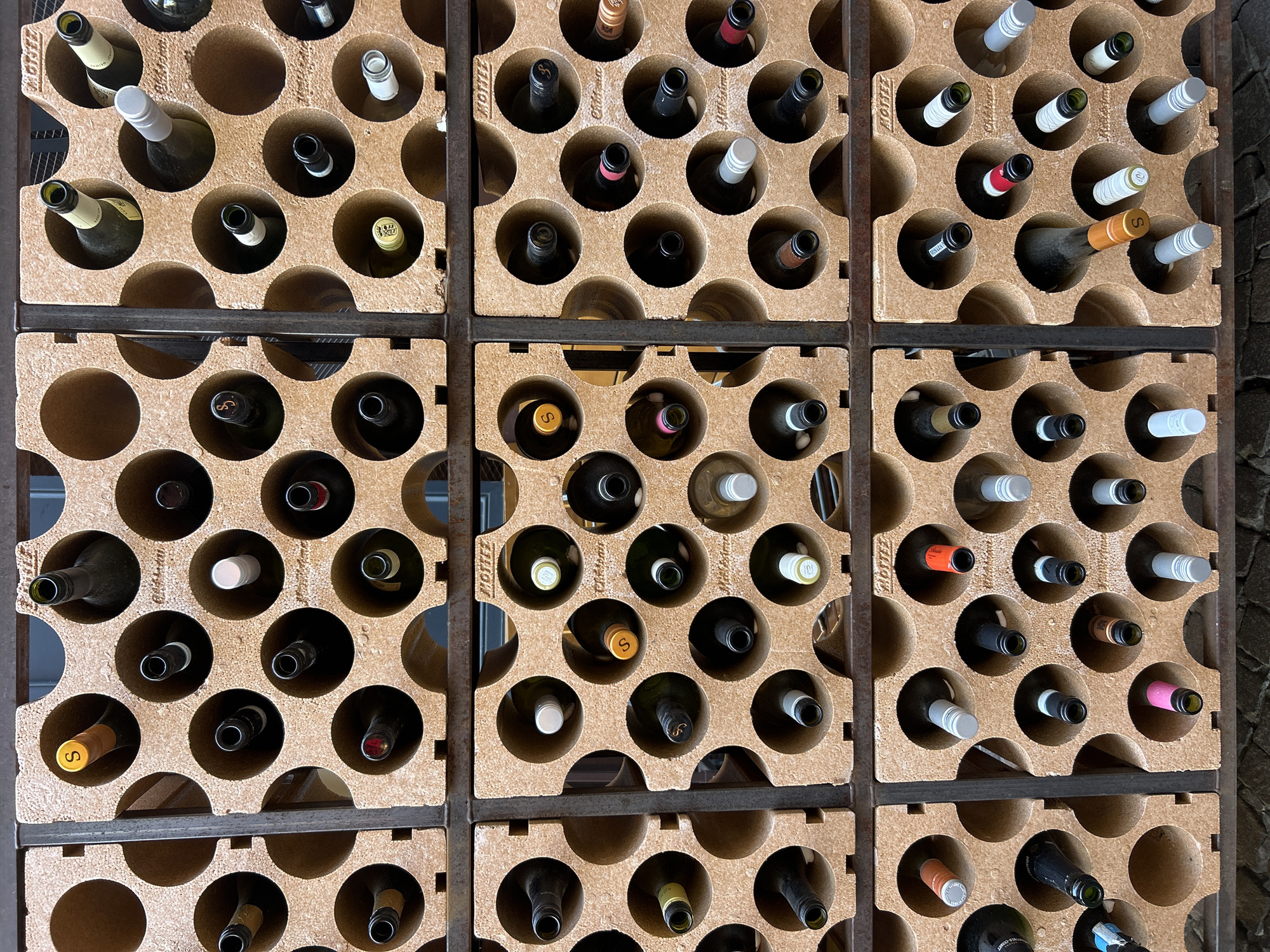 The image shows a close-up of a wine rack filled with wine bottles. The rack is made of a textured, light brown material with circular openings for each bottle. The bottles are arranged in rows and columns, with many of them partially visible. Some bottles have visible labels or caps of various colors, including yellow, white, red, and silver. The overall setting appears to be indoors, with a rustic or cellar-like aesthetic. The wine rack is a modular unit, with multiple such units visible, suggesting a larger storage or display area. Based on the presence of the text "Mottez" visible on the rack in multiple locations, this is likely a Mottez wine rack. Information available in the public domain indicates that Mottez wine racks are often found in establishments selling or storing wine. Without additional contextual information or visible landmarks within the image itself, it is not possible to definitively pinpoint the specific location in Leuven, Belgium. However, if this rack is part of a commercial establishment, it could be found in a wine shop, restaurant, or cellar in the city.