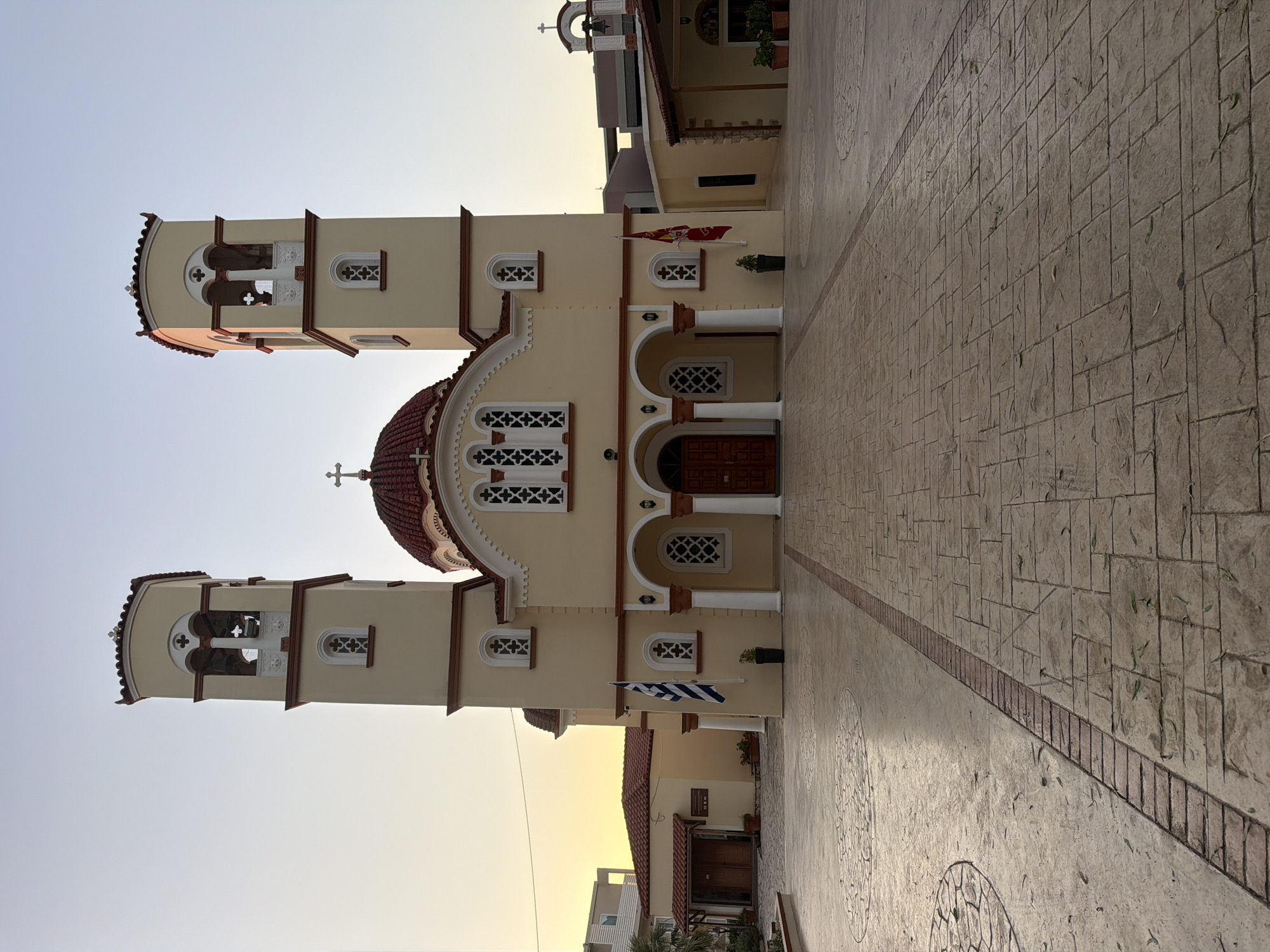The image shows the exterior of the Holy Church of the Assumption of the Virgin Mary in Georgioupoli, Crete, Greece. The church is a traditional Greek Orthodox church with a yellow facade, a red-tiled dome, and two bell towers. The bell towers are square with arched openings for bells, and each is topped with a small cross. The main entrance to the church features a portico supported by white columns and large wooden double doors. Above the entrance, there are several arched windows with intricate white lattice work. Two flags are displayed on the facade: a Greek flag on the left and another flag on the right. The church is situated in a paved courtyard with patterned stonework and decorative circular inlays. In the background, there are some low-rise buildings and trees. The sky is clear and light, suggesting it is either early morning or late afternoon. The scene is tranquil and devoid of any people.