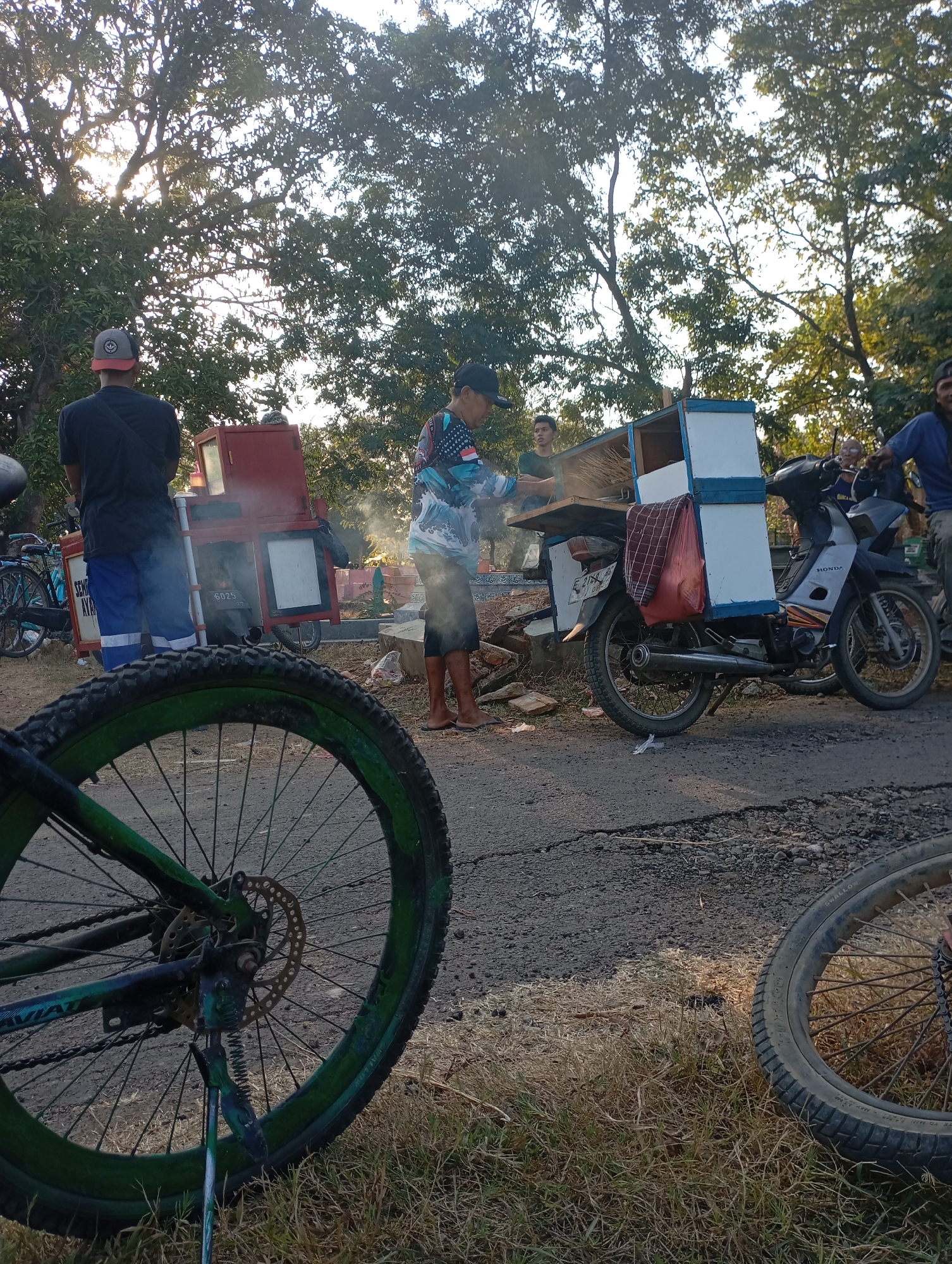 In Kecamatan Haurgeulis, Indonesia, on a paved road shaded by trees with dappled sunlight, a group of people are gathered. In the foreground, the wheel of a bicycle, painted green and black, is prominently displayed. To the left, another bicycle is parked, its rear wheel visible.
In the mid-ground, a vendor is preparing food. He stands behind a mobile cart, which is attached to a motorcycle. The cart has a white and blue structure on top, with a red section below containing a glass display. A red and white checkered cloth hangs from the side of the cart. Smoke emanates from the cart, obscuring the vendor's lower body. The motorcycle, a Honda, is parked to the right of the cart, its wheels and body visible.
Several other individuals are present. One man, wearing a black shirt and blue pants, stands to the left of the cart, facing away from the camera. Another person, in a blue shirt and a cap, is seated on the motorcycle. A younger person, wearing a dark green shirt, stands behind the vendor. The background shows more trees and what appears to be a cemetery with concrete structures. The overall scene depicts a busy street-side vending operation.