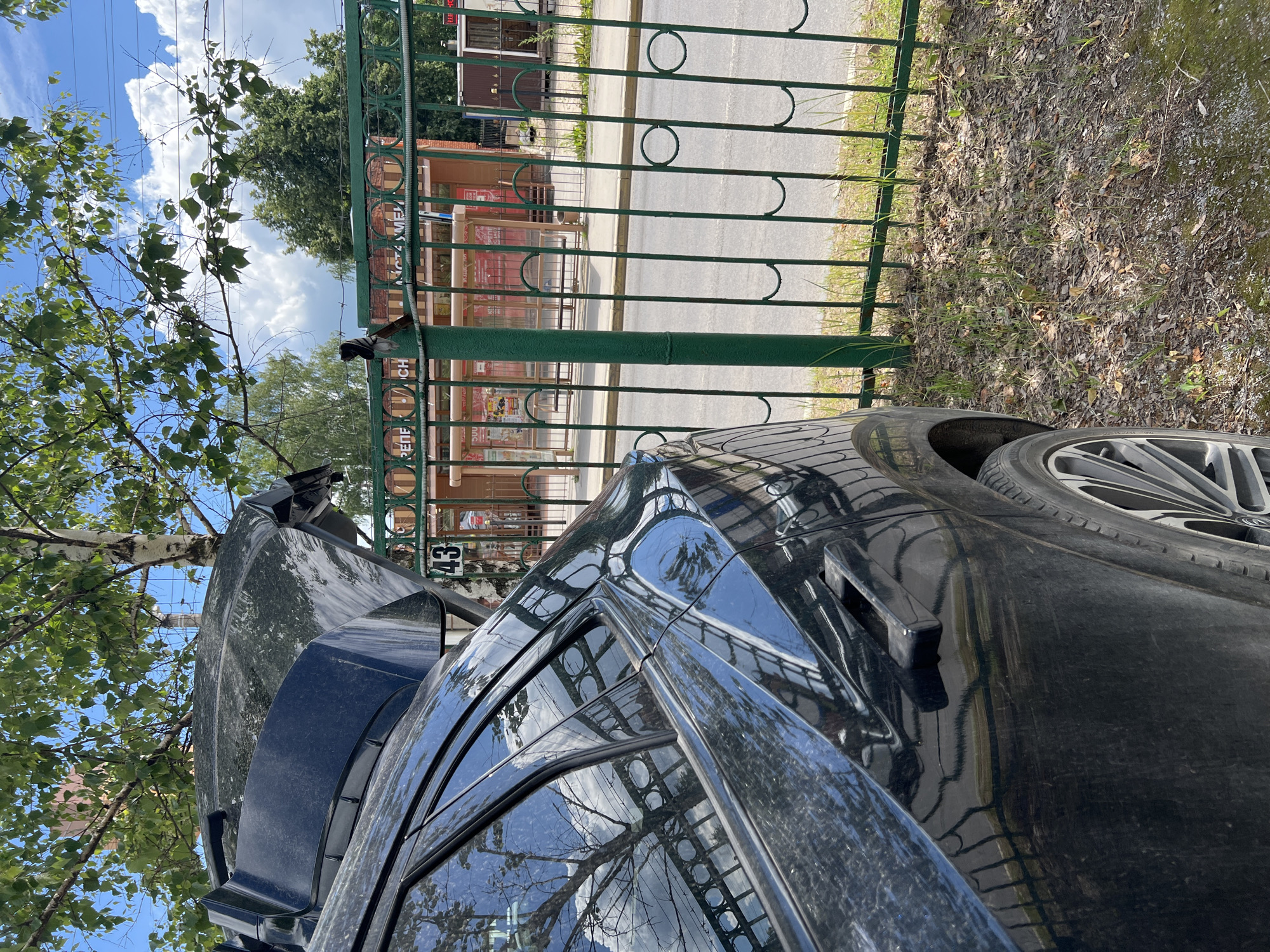 A black SUV with its trunk open is parked next to a green metal fence. The fence has a decorative pattern of circles and arches. Behind the fence, there is a bus stop shelter with advertisements visible. In the background, trees and buildings can be seen under a blue sky with white clouds. The license plate number on the fence is "43". The scene appears to be on a street in Tula, Russia, possibly near a residential area or a public transportation stop. The ground is covered with gravel, leaves, and sparse grass.