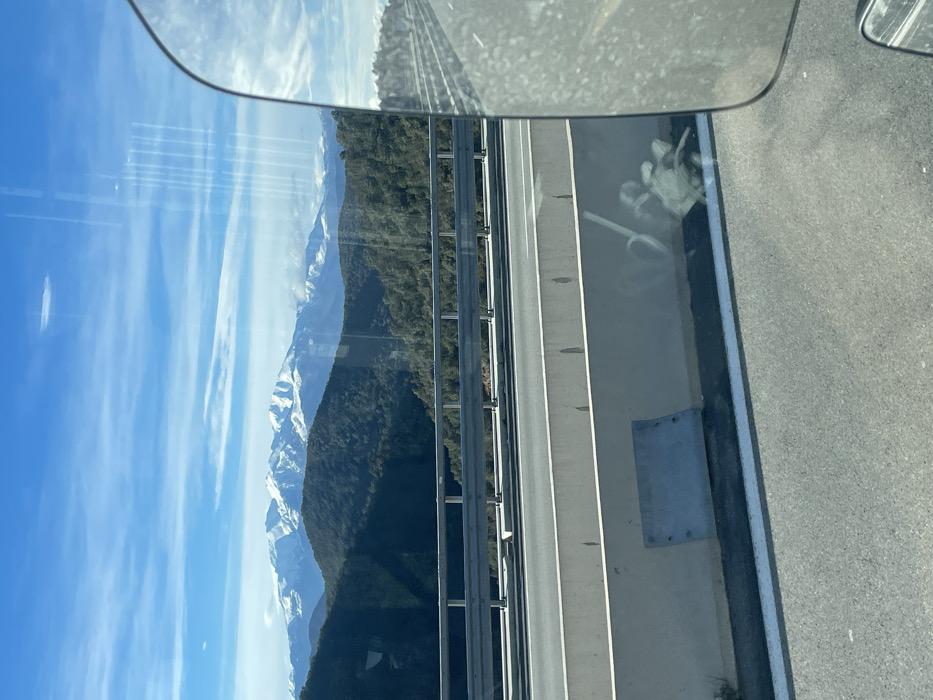 The image depicts a panoramic view from a moving vehicle on a multi-lane highway near Le Perthus, France, looking towards a mountainous landscape under a clear blue sky. In the immediate foreground, to the left, a light-colored concrete highway barrier is visible, featuring a rectangular metal plate bolted near its base. Above this barrier, a silver metal guardrail runs horizontally. The asphalt highway surface is marked with white lines, and reflections of vertical streaks and faint shapes are apparent on the window through which the photograph was taken. A significant portion of a vehicle's side mirror, likely a truck or bus, occupies the right side of the frame, reflecting a distorted view of the road and sky. Beyond the highway infrastructure, a valley with densely forested, dark green hills extends into the midground. In the far background, majestic snow-capped mountain ranges rise prominently, contrasting with the blue sky that contains sparse white clouds. No people or overt actions are visible; the scene captures a static landscape view from a dynamic perspective.